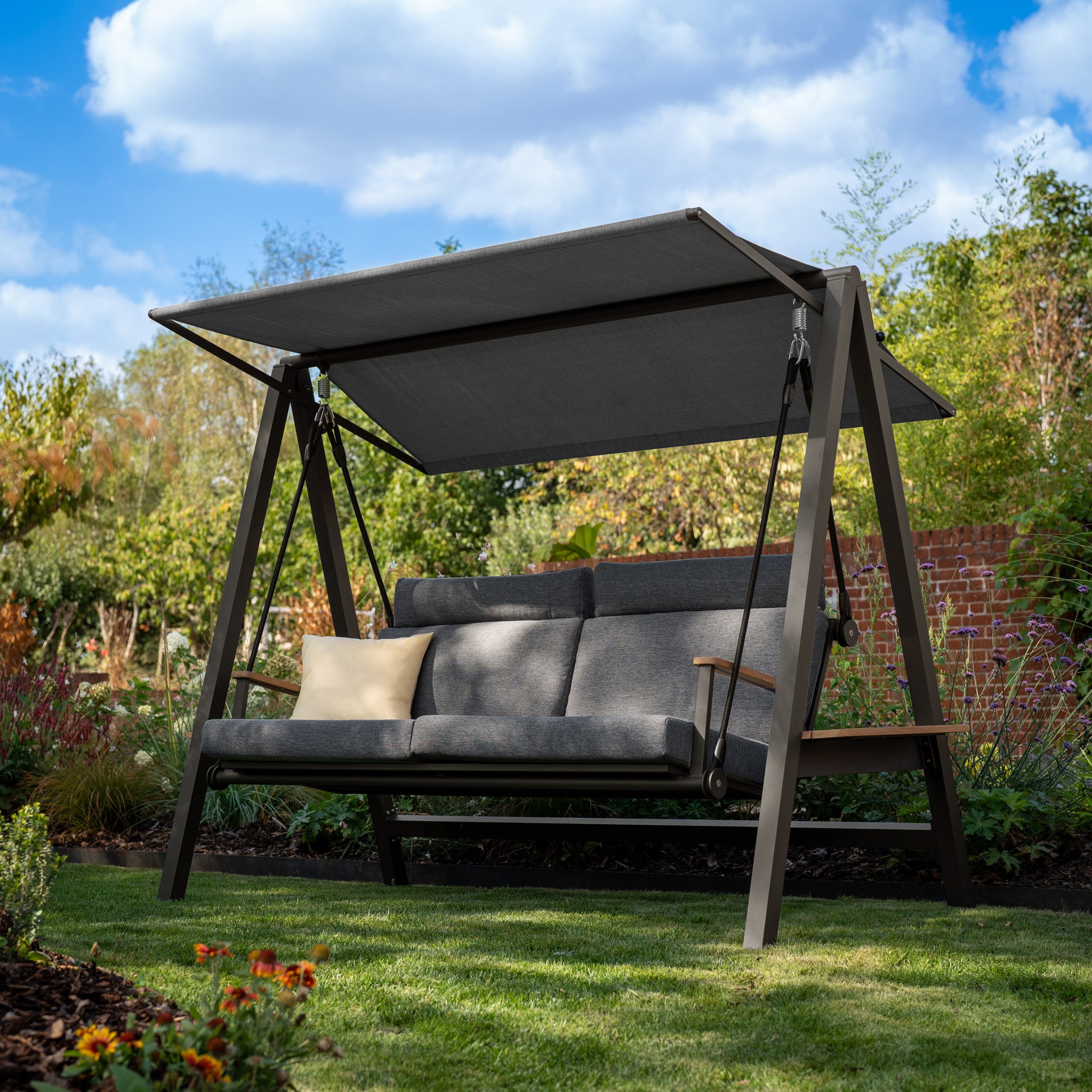 The Lima Deluxe Aluminium Swing Seat in Charcoal showcases an outdoor swing bench with grey cushions and a robust olefin canopy, set amidst a grassy area adorned with trees and flowers. Its powder-coated aluminium frame guarantees lasting elegance and stability.