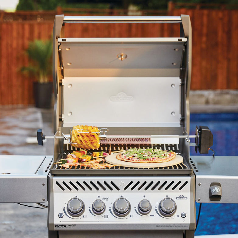 A Napoleon Rogue XT 425 SIB Gas Barbecue with a stainless steel finish showcases vegetables, chicken skewers, and a pizza cooking on it, all while an inviting pool and wooden fence enhance the picturesque backdrop.