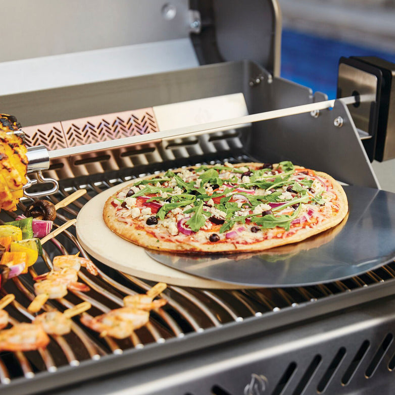 A pizza topped with vegetables and arugula bakes on a Napoleon Rogue SE 425 RSIB Gas Barbecue, while kebabs sizzle on the infrared side burner and grilled corn accompanies them.