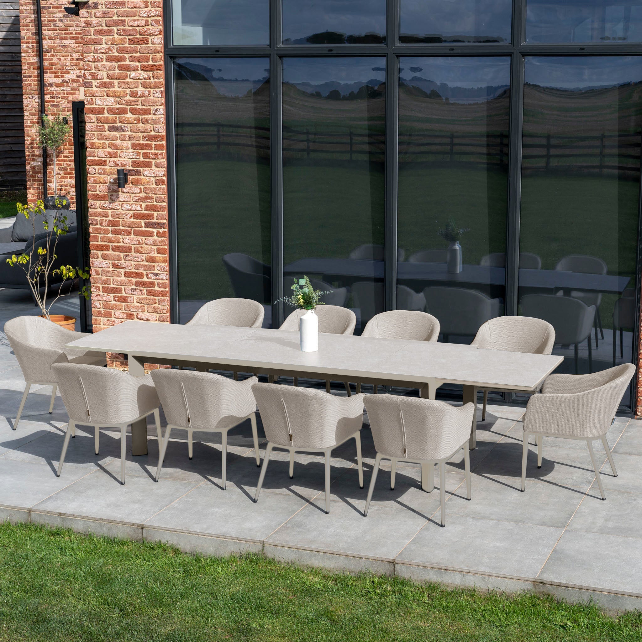 Outdoor dining area showcasing the Luna 10 Seat Outdoor Fabric Extending Ceramic Dining Set in Fawn, complete with weather-resistant chairs and a decorative vase elegantly positioned on the table, all set against the backdrop of glass windows on the patio.