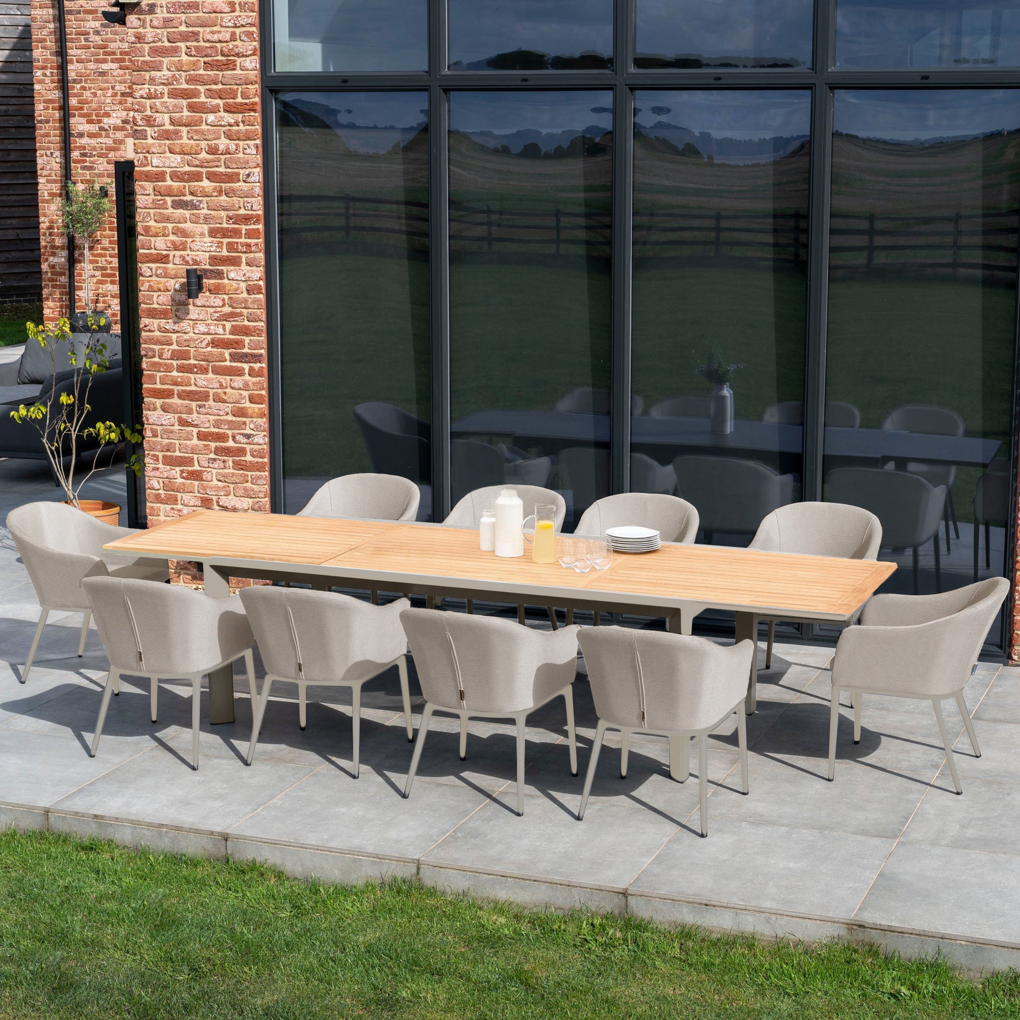 An inviting outdoor patio setup showcases the Luna 10 Seat Outdoor Fabric Extending Teak Dining Set in Fawn, with chairs made from all-weather materials placed elegantly on a stone terrace near expansive glass windows.