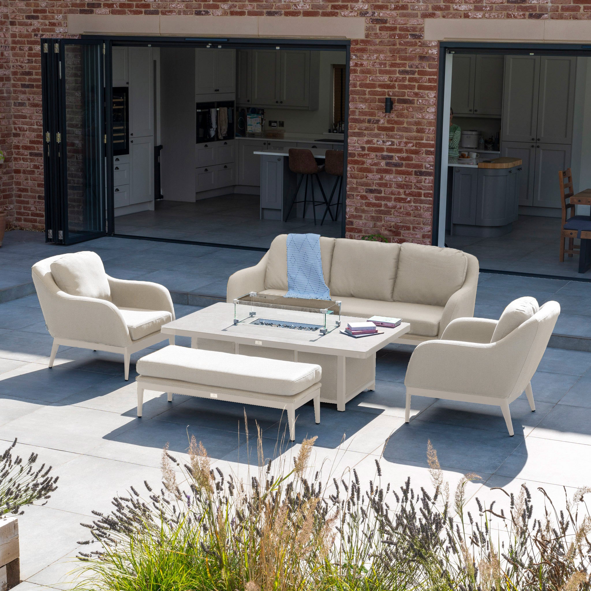 Outdoor patio featuring the Luna 3 Seat Outdoor Fabric Sofa Set in Fawn, crafted from all-weather material, set on a tile floor near a brick house with open sliding doors, complemented by a rising firepit table.