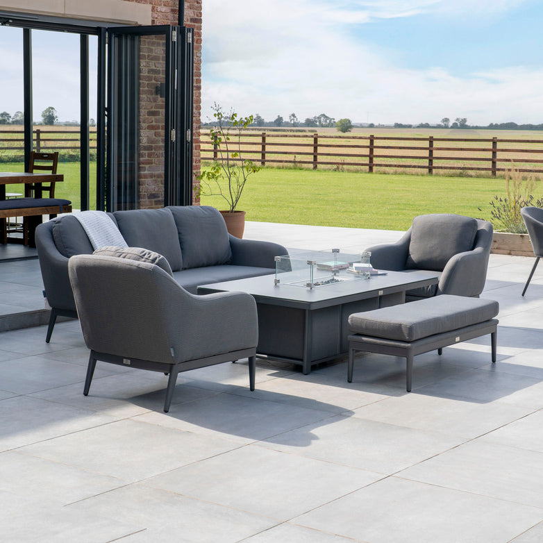 Outdoor patio set featuring the Luna 3 Seat Outdoor Fabric Sofa Set with Rising Firepit Table in Grey, complemented by a coffee table and gray cushions, all against the scenic view of a green field with a wooden fence.