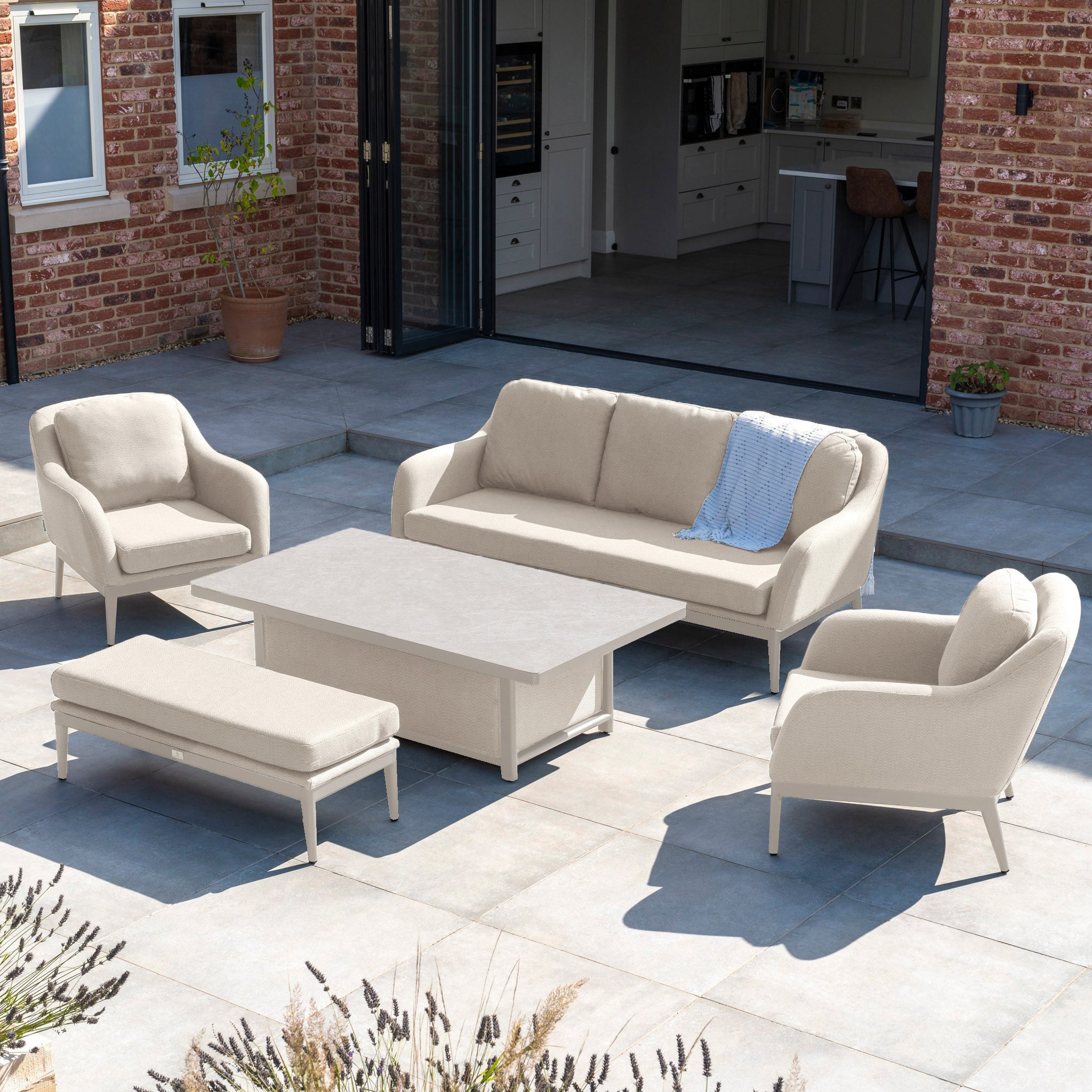 The outdoor patio features the Luna 3 Seat Outdoor Fabric Sofa Set with Rising Table in Fawn, elegantly positioned on gray stone tiles. This maintenance-free furniture set includes a beige sofa, two armchairs, a bench, and a table. Made from all-weather materials, it offers the perfect spot to unwind by the brick house with minimal upkeep required.