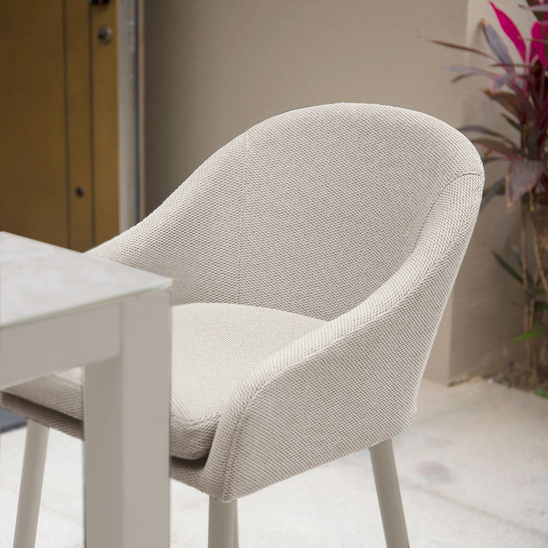 A Luna Outdoor Fabric Bar Stool in Fawn, crafted from weather-resistant outdoor fabric, sits next to a table on a patio, near a wall with a potted plant.