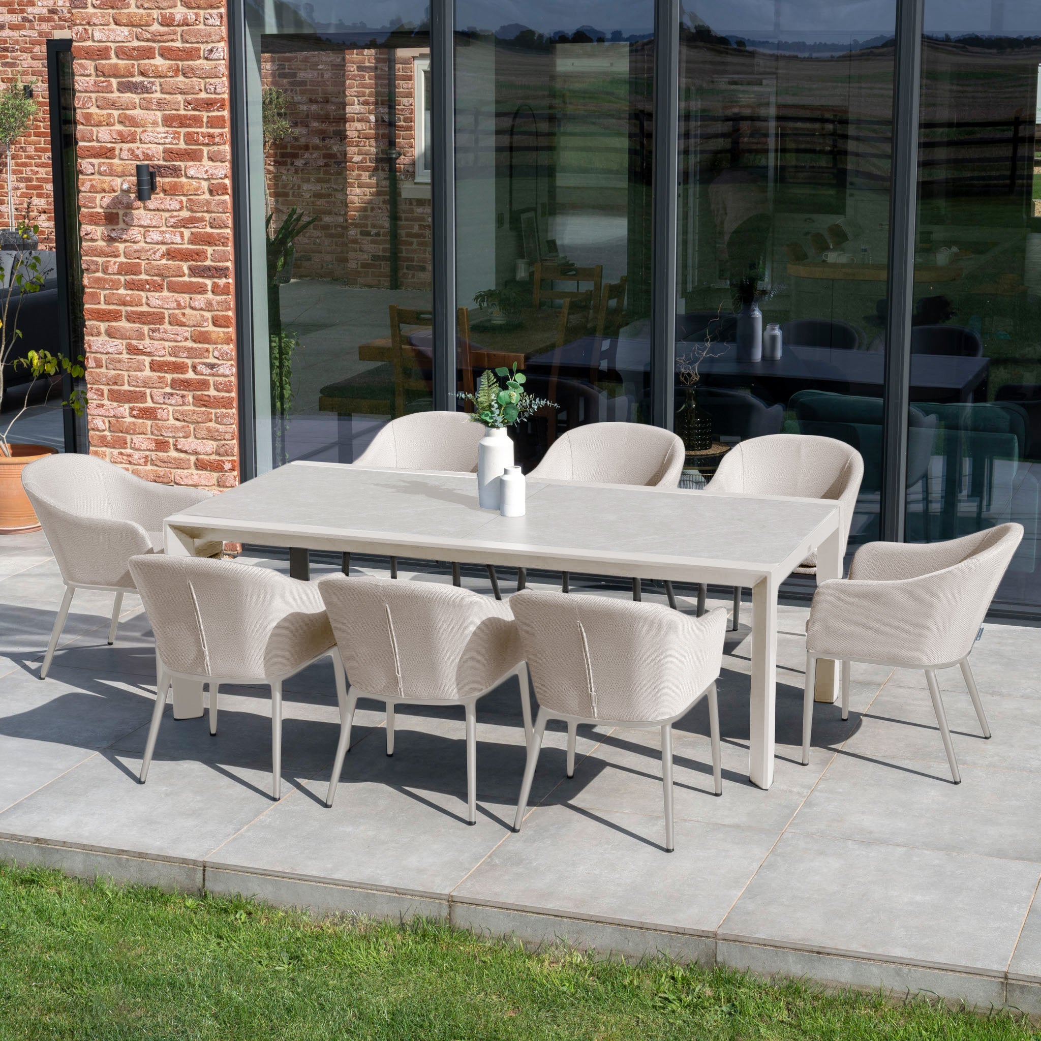 The Luna 8 Seat Outdoor Fabric Extending Ceramic Dining Set in Fawn enhances this dining arrangement, showcasing eight fawn chairs surrounding a rectangular table on the patio by the large glass doors. Its all-weather material guarantees durability and elegance throughout the seasons.