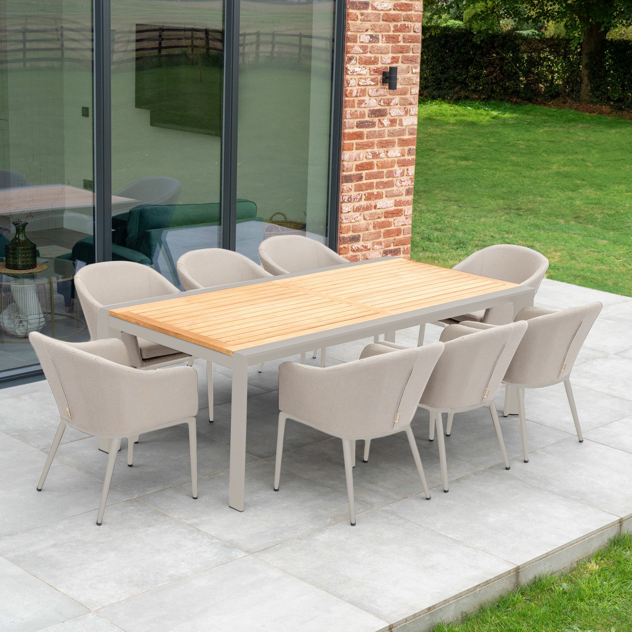 The Luna 8 Seat Outdoor Fabric Extending Teak Dining Set in Fawn enhances the patio beside a brick wall and glass door, all made from all-weather material, overlooking a lush grassy lawn.