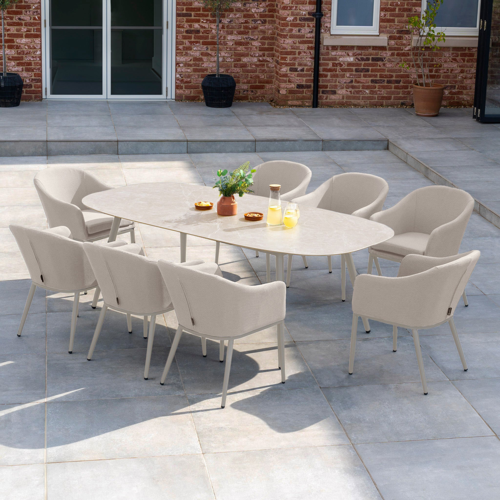 Experience outdoor dining with the Luna 8 Seat Outdoor Fabric Oval Ceramic Dining Set in Fawn, featuring a chic ceramic table, eight weather-resistant chairs in complementary tones, potted plants, and refreshing beverages on the patio.