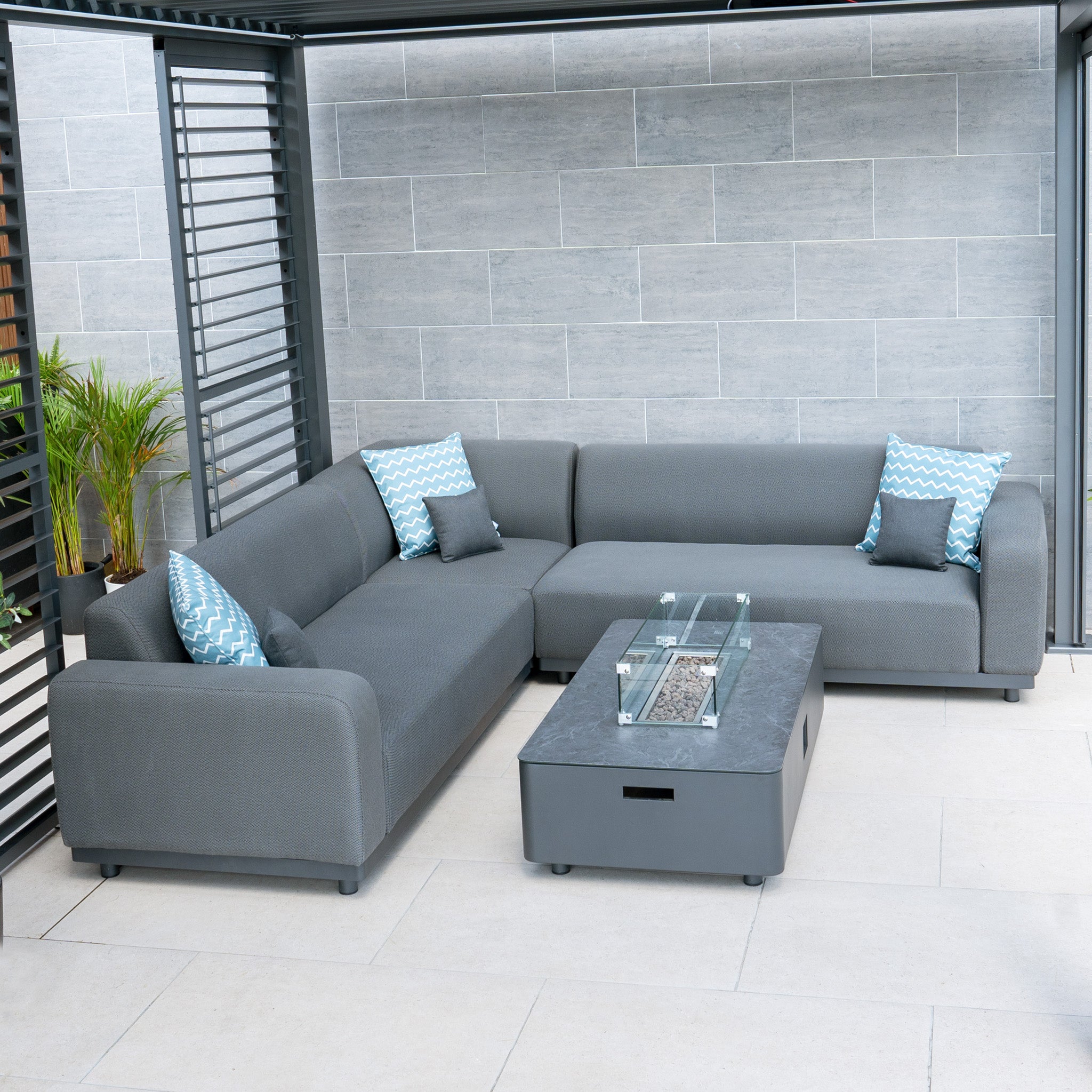 Modern outdoor patio featuring the Luna Outdoor Fabric Corner Sofa Set with a durable gray sectional, blue patterned pillows, and a maintenance-free rectangular glass-top firepit coffee table.