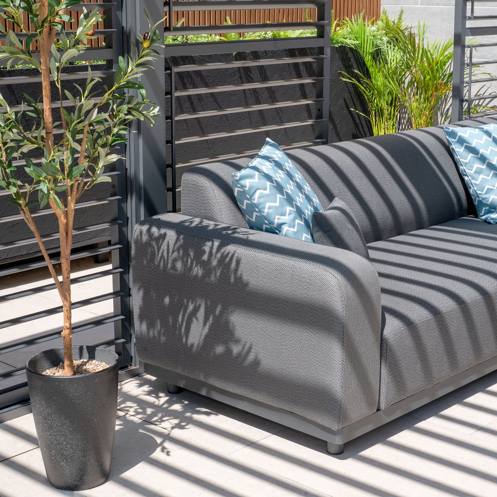 A modern patio showcases a Luna Outdoor Fabric Corner Sofa Set with Coffee Table in grey, adorned with blue-patterned pillows and a potted plant casting shadows in the sunlight. The all-weather material offers durable, stain-proof comfort.