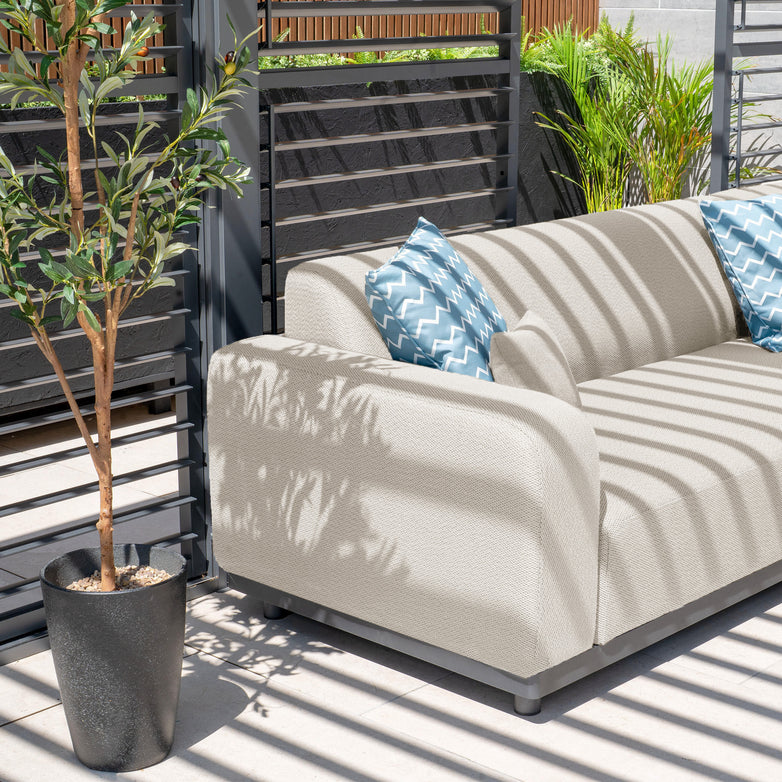 Relax in style with the Luna Outdoor Fabric Corner Sofa Set in fawn, featuring all-weather material and white fabric. Accented by two blue-patterned pillows and a potted tree, it creates a serene space under dappled sunlight for a maintenance-free oasis.