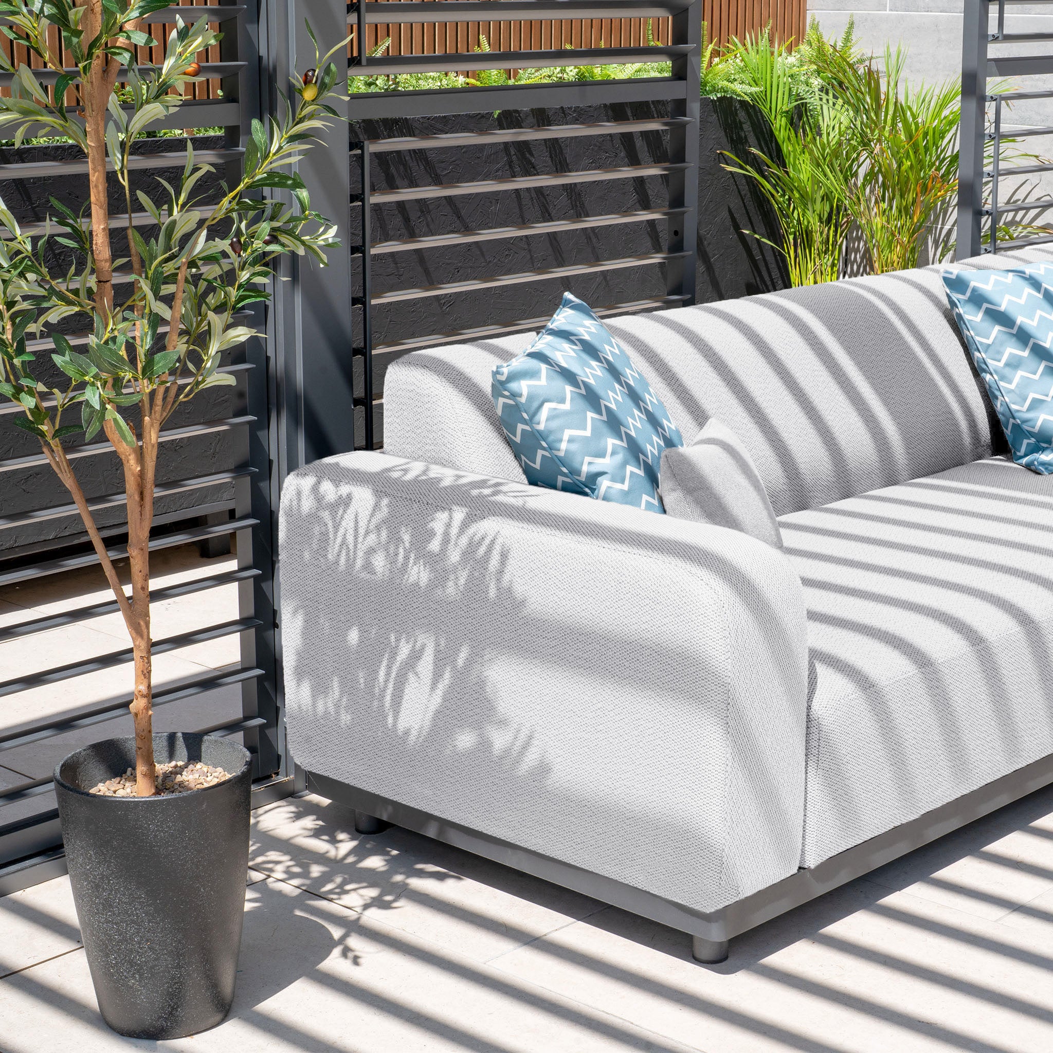 The Luna Outdoor Fabric Corner Sofa Set with Coffee Table in Oyster Grey features an all-weather sofa adorned with blue patterned pillows. It rests elegantly under a slatted pergola, accompanied by a nearby potted plant.