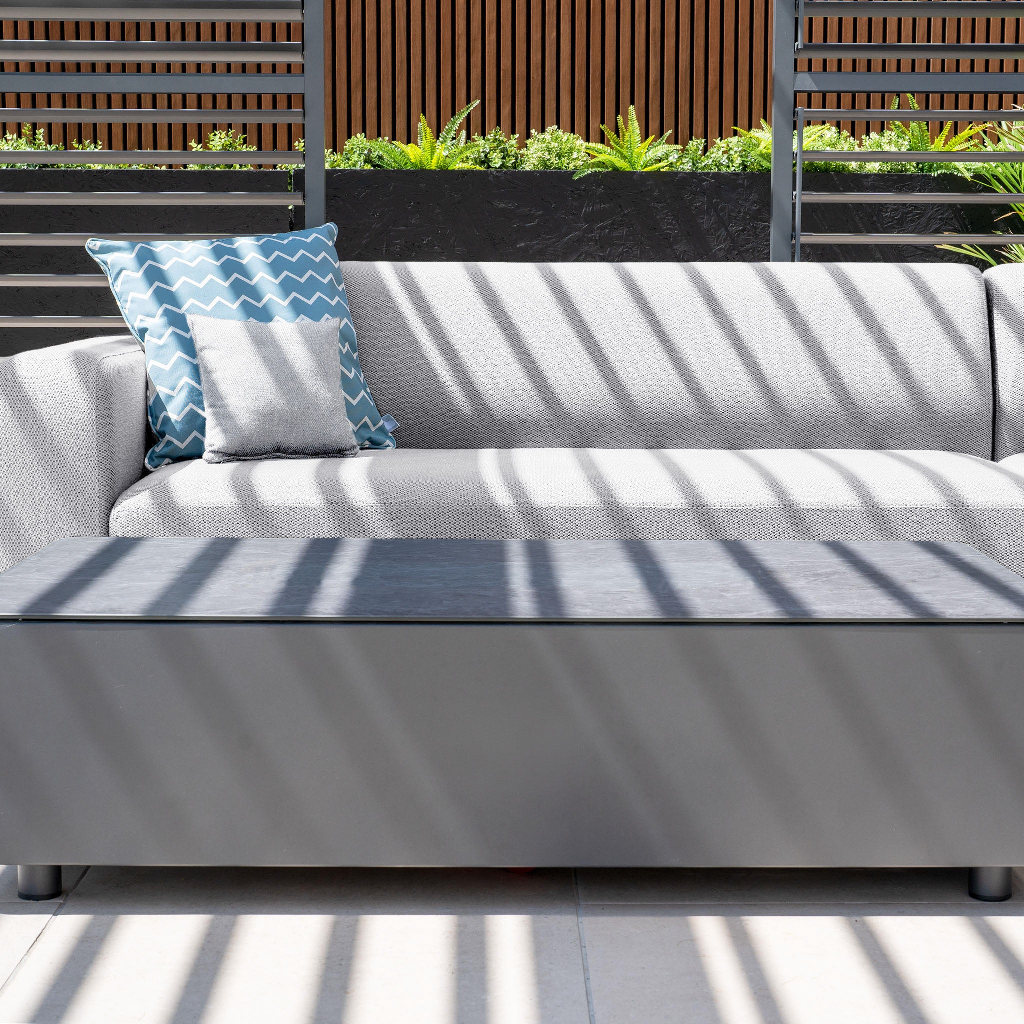 The Luna Outdoor Fabric Corner Sofa Set in Oyster Grey with a black coffee table and two pillows, including a zigzag patterned one, enjoys striped shadows under sunlight. Made from all-weather materials, it’s ideal for any season.