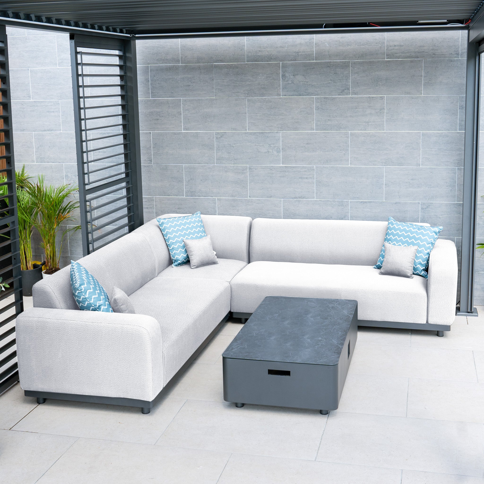 Outdoor patio featuring the Luna Outdoor Fabric Corner Sofa Set in Oyster Grey, with an L-shaped sofa, blue-patterned pillows, and a black coffee table. This all-weather set is perfect for enduring the elements against gray tiled walls.