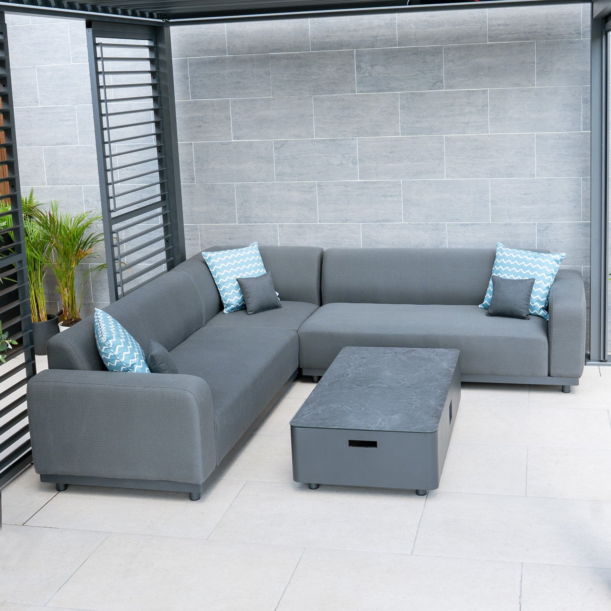 A stylish outdoor seating area features the Luna Outdoor Fabric Corner Sofa Set with Coffee Table in Grey, complete with blue patterned pillows. This setup provides comfort and durability with stain-proof, all-weather material.