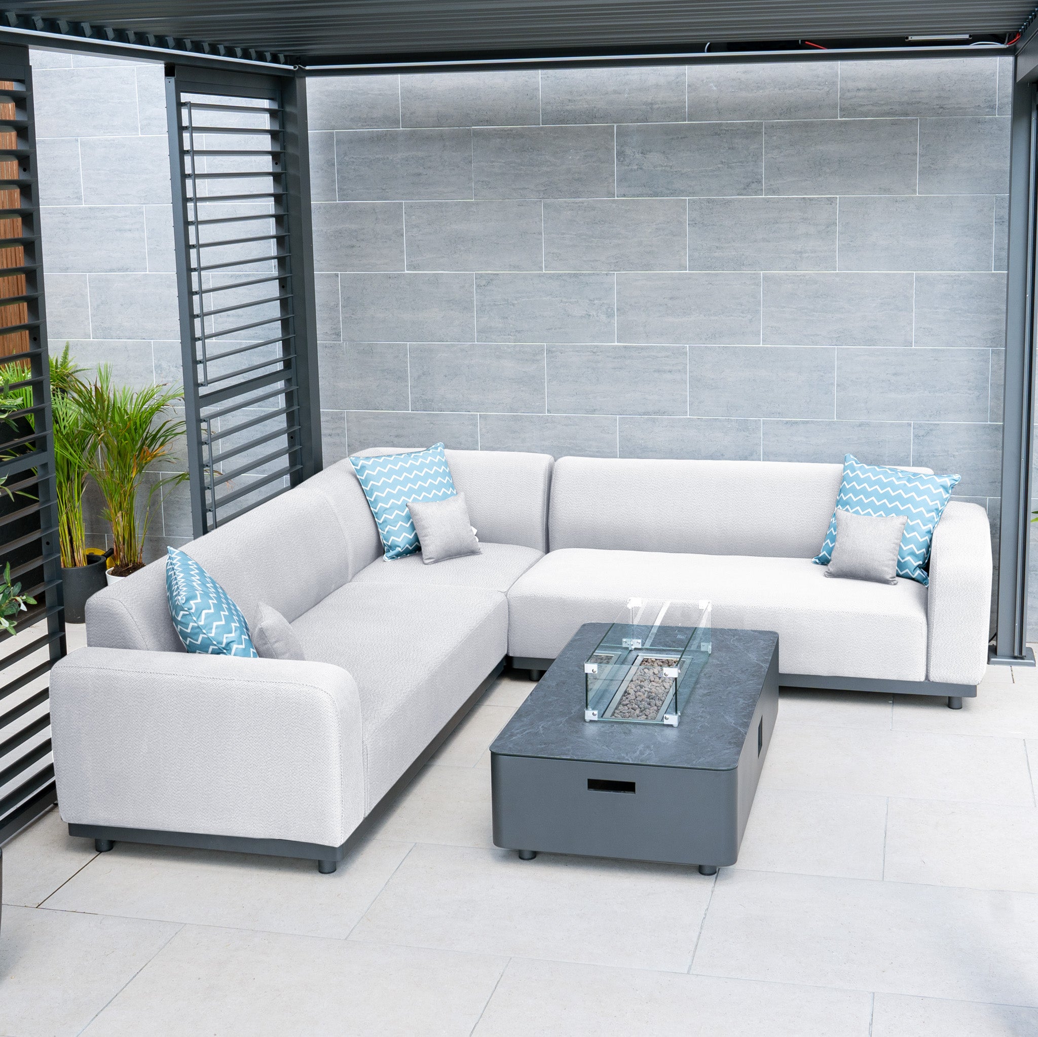 A stylish patio showcases the Luna Outdoor Fabric Corner Sofa Set in Oyster Grey, featuring blue-patterned pillows and enhanced with a Harbour Lifestyle firepit coffee table for added elegance and durability.