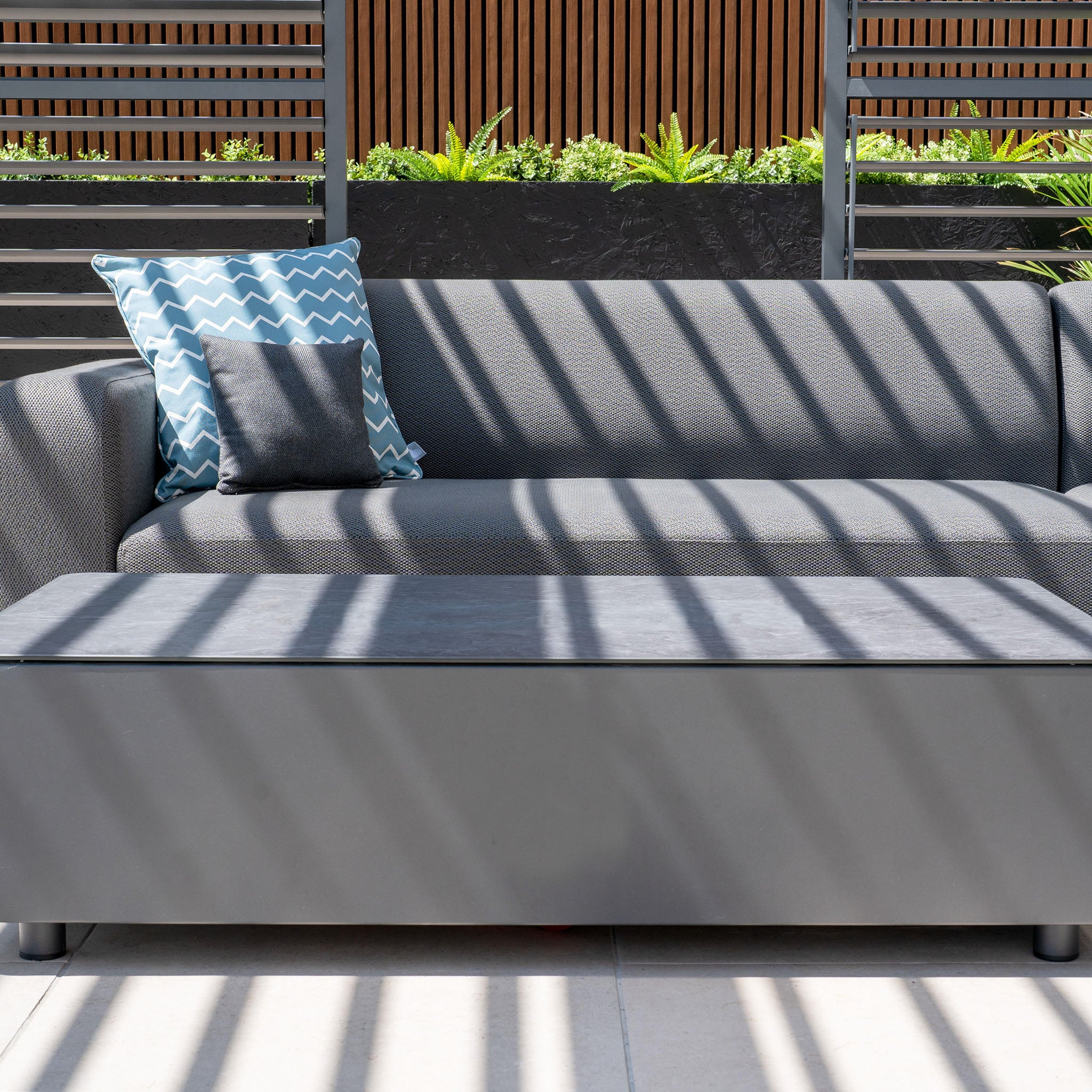 The Luna Outdoor Fabric Corner Sofa Set in Gray includes a stain-proof sofa with two cushions (one patterned) framed by striped shadows, and a matching coffee table.