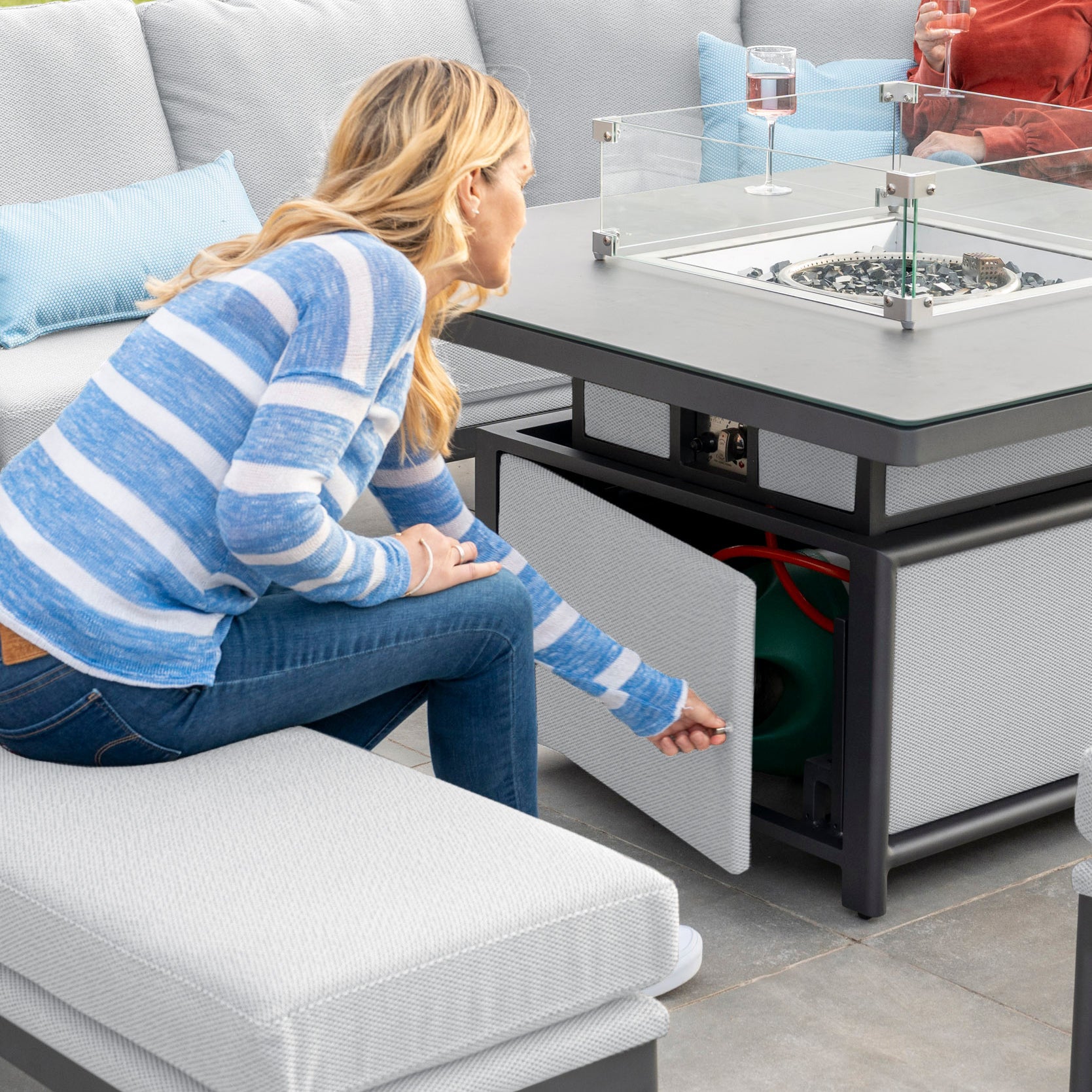 A person in a blue striped sweater opens a cabinet under the Rising Firepit Table, with friends seated nearby on the Luna Deluxe Outdoor Fabric Square Corner Dining Set in Oyster Grey.