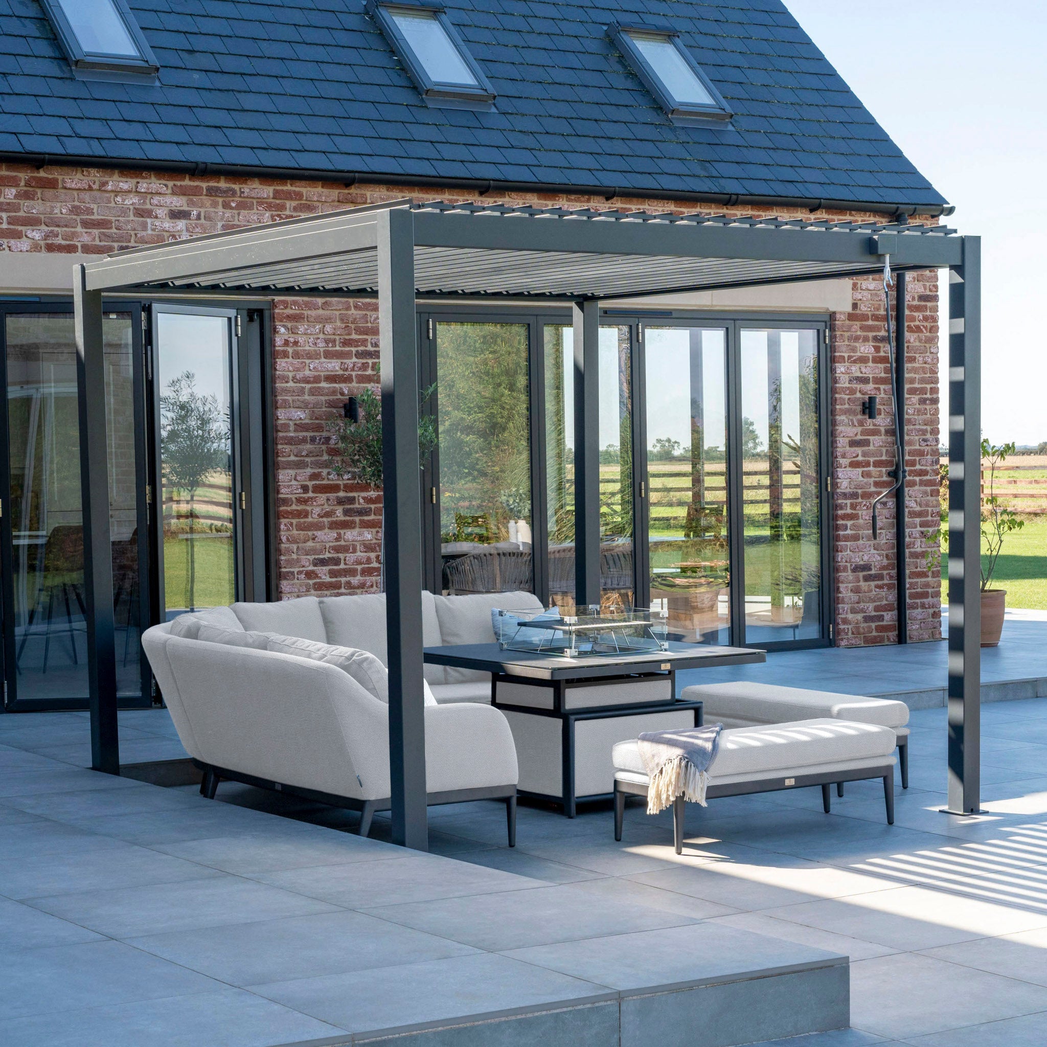 Modern outdoor patio with a pergola, the Luna Deluxe Outdoor Fabric Square Corner Dining Set with Rising Firepit Table in Oyster Grey, and an ottoman in a brick house setting.