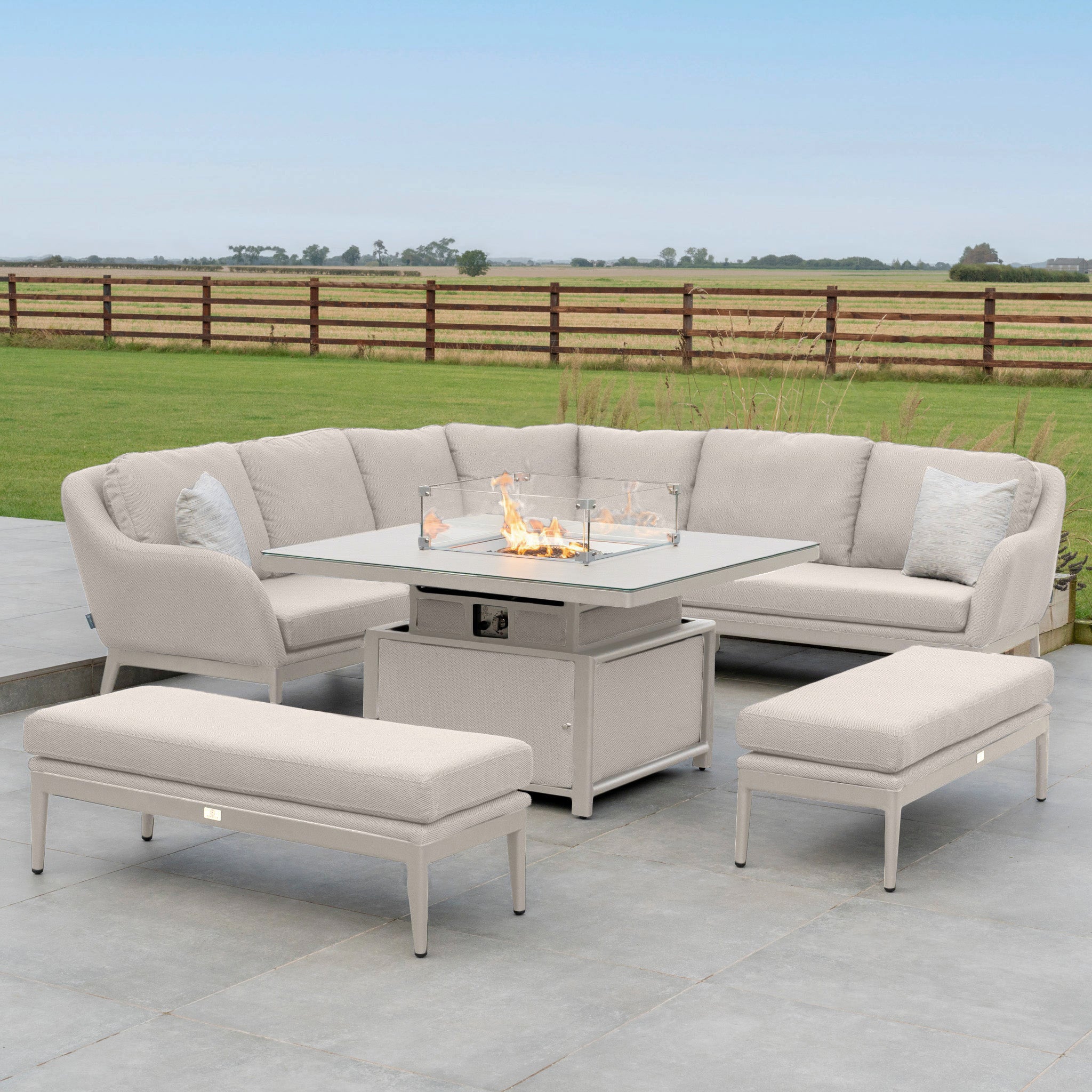 The outdoor patio features the Luna Deluxe Outdoor Fabric Square Corner Dining Set in Fawn, made from stain-proof fabric, complete with a rising firepit table set on a stone surface. The arrangement overlooks a picturesque field and fence.