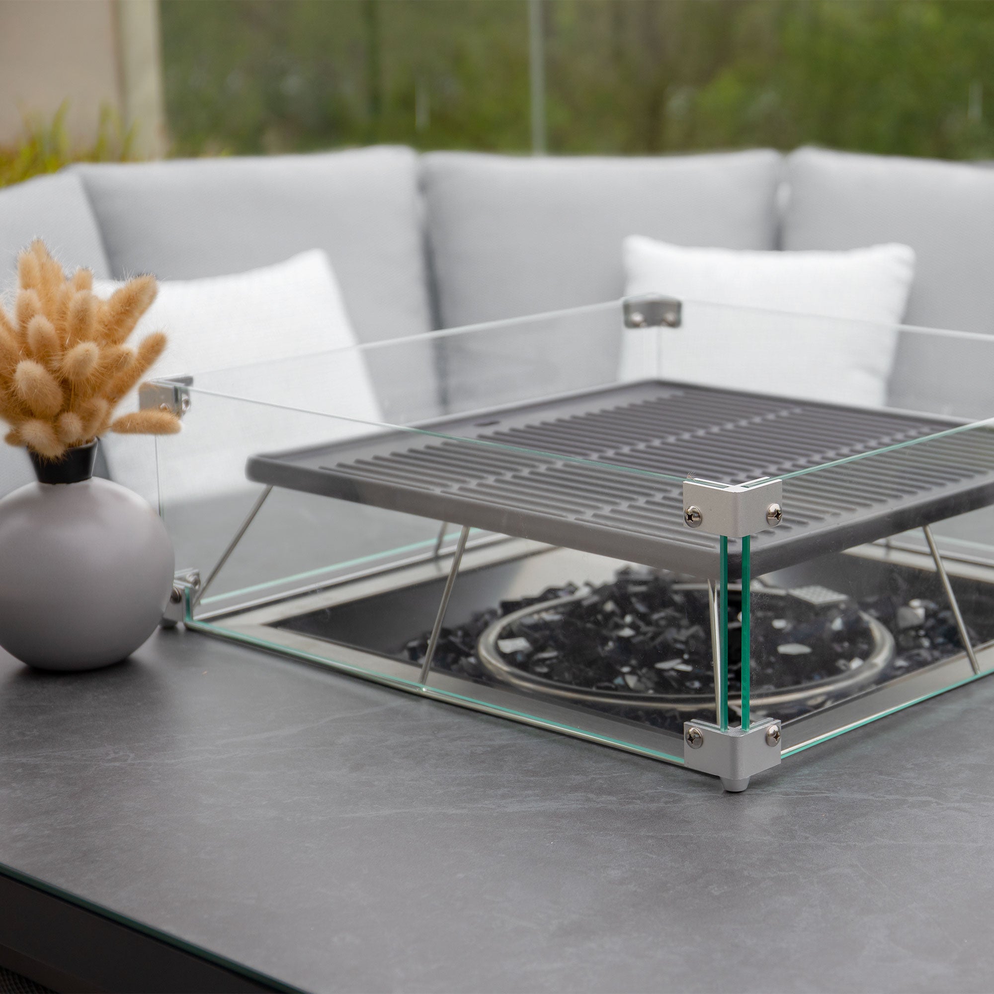 Close-up of a modern tabletop gas fire pit with glass panels, gray cushions in the background, and a small vase of dried flowers. The sleek design perfectly complements the Luna Deluxe Outdoor Fabric Square Corner Dining Set with Rising Firepit Table in Oyster Grey, creating an inviting atmosphere.