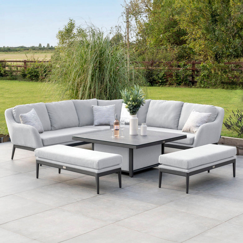 Enjoy your outdoor space with the Luna Deluxe Outdoor Fabric Square Corner Dining Set, featuring a grey cushioned L-shaped sofa, two benches, and an adjustable table with a stylish vase. Crafted from maintenance-free materials and adorned in elegant Oyster Grey fabric, this set perfectly complements a tiled patio by a lush green lawn.