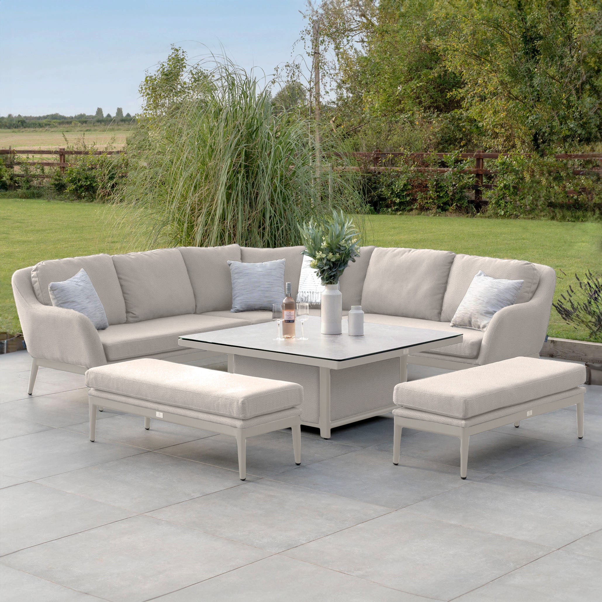 An outdoor patio showcasing the Luna Deluxe Outdoor Fabric Square Corner Dining Set with Rising Table in Fawn, featuring an L-shaped sofa, cushioned benches, and a vase-topped rising table. The stain-proof design beautifully contrasts with the vibrant green landscape in the background.