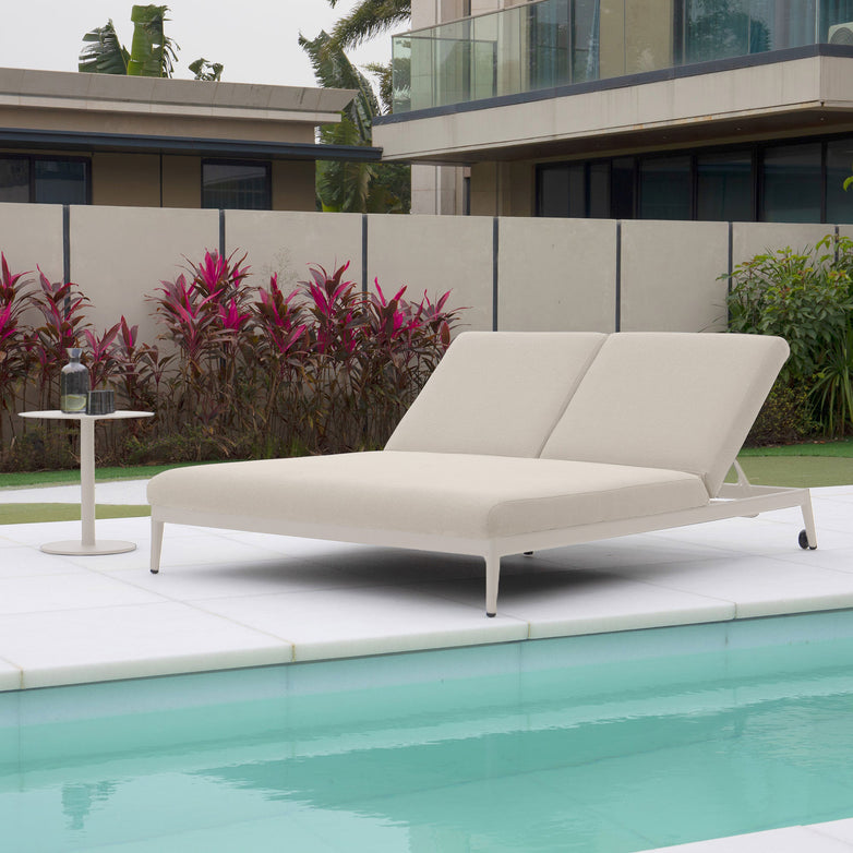 The Luna Outdoor Fabric Double Sun Lounger in Fawn, designed for all-weather conditions, is paired with a small table featuring a bottle and glass, set amidst lush tropical plants.