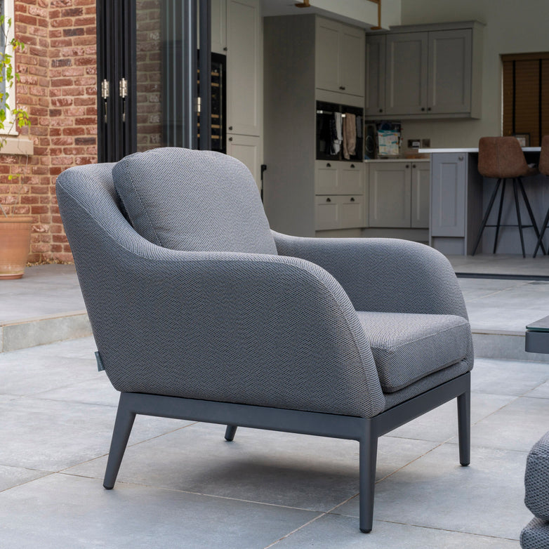 A Luna Armchair in Grey, crafted by Harbour Lifestyle and featuring comfortable outdoor fabric, is placed on a patio before an open sliding door that opens into a modern kitchen with light gray cabinets.