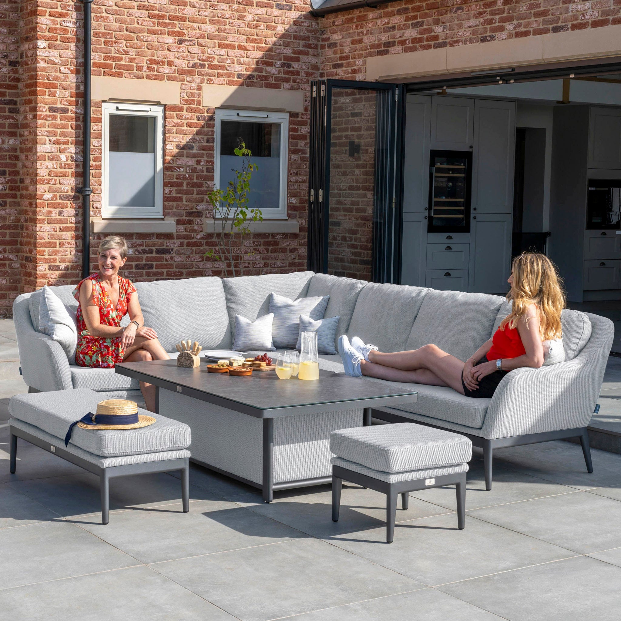 Two women relax on an outdoor patio set made with the Luna Outdoor Fabric Rectangular Corner Dining Set with Rising Table in Oyster Grey (Left Hand), enjoying drinks and snacks laid out on the maintenance-free, durable dining set in front of them.