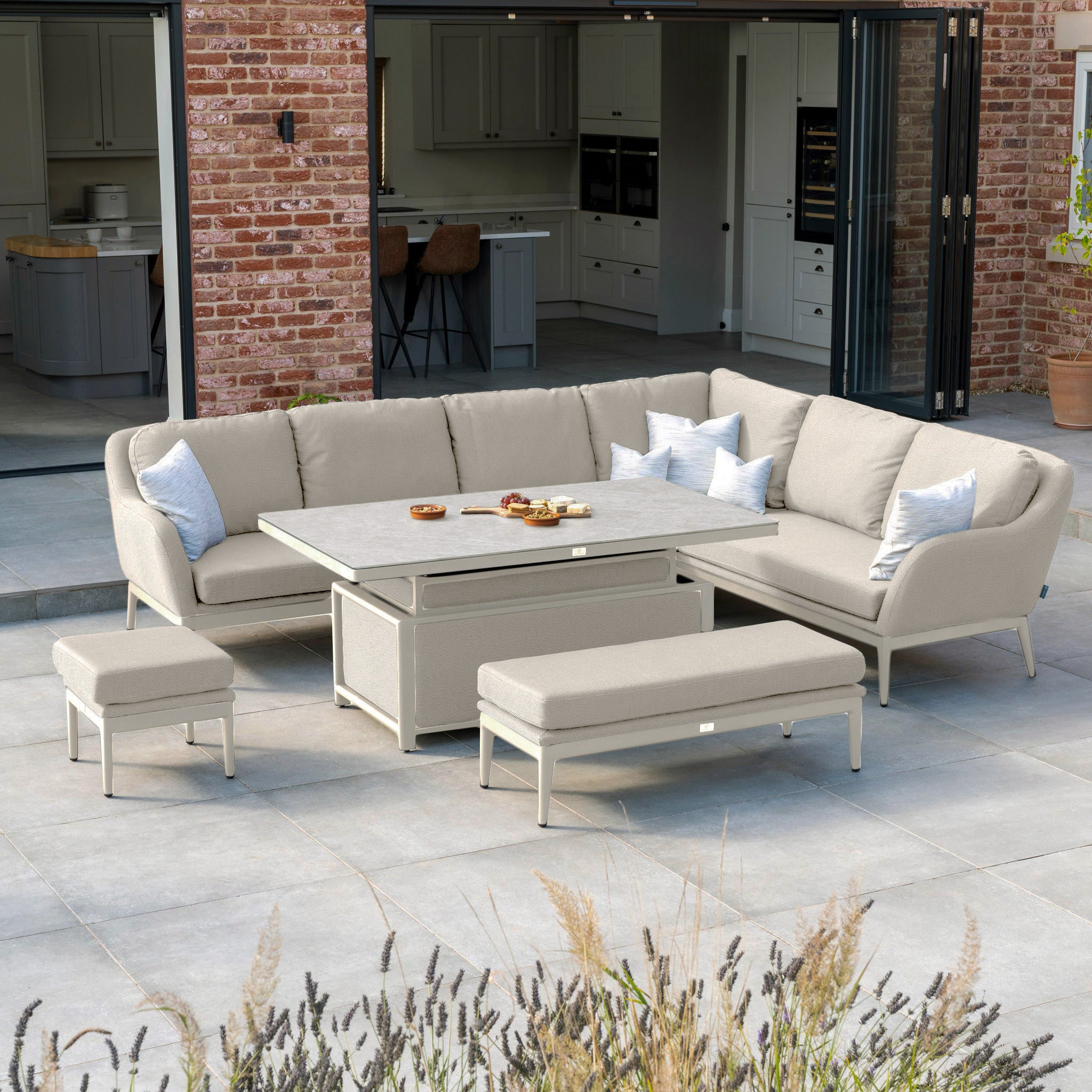 A patio is adorned with the Luna Outdoor Fabric Rectangular Corner Dining Set in Fawn (Right Hand), which includes a beige sectional sofa and stools encircling an adjustable, maintenance-free table, while open doors invite you into the indoor kitchen.