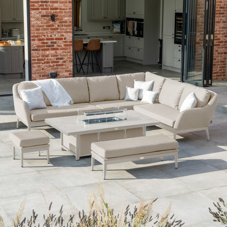 A contemporary outdoor space featuring the Luna Outdoor Fabric Rectangular Corner Dining Set in Fawn with a right-hand layout. The sectional sofa includes cushions made of outdoor fabric, and the set is completed with benches resting on a tiled floor. A rising firepit table enhances both warmth and style against the brick facade.