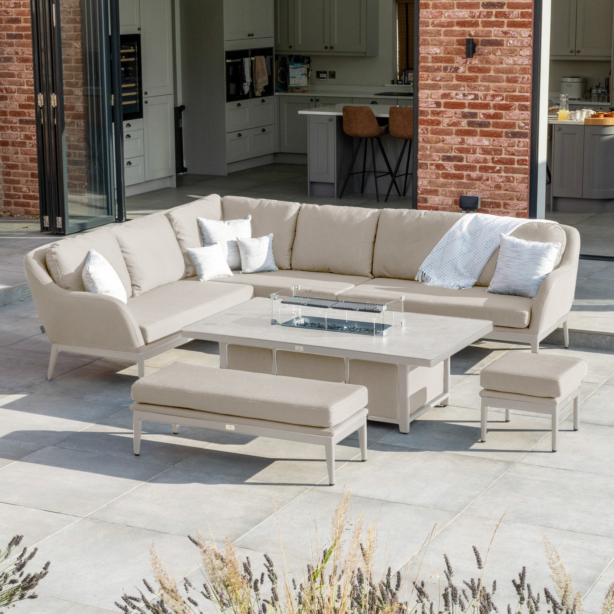 The Luna Outdoor Fabric Rectangular Corner Dining Set with Rising Firepit Table in Fawn (Left Hand) showcases a modern style with its rising firepit table and beige cushions, situated on a tiled patio near open glass doors. It is expertly crafted from all-weather materials and durable outdoor fabric.