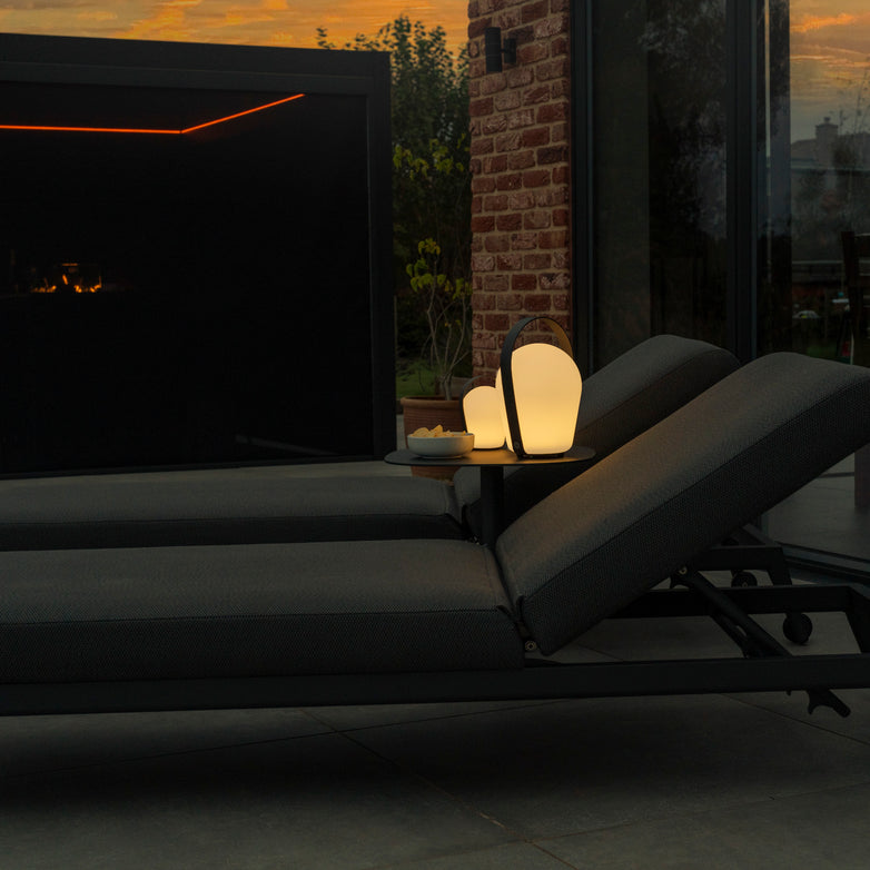 Two lounge chairs with glowing Bob Large Table Lights in Charcoal and weather-resistant lanterns near a modern brick wall and patio area at sunset.