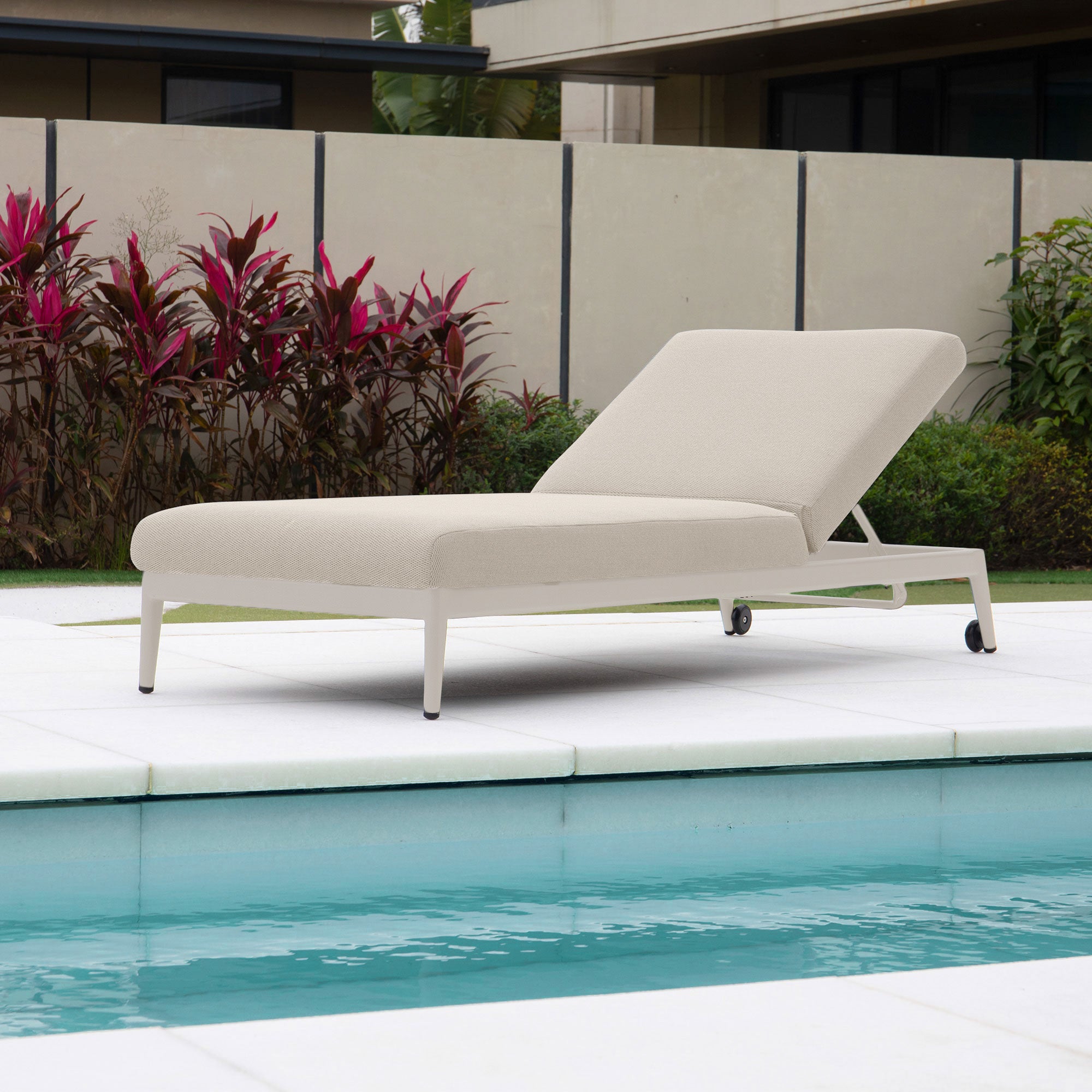 The Luna Outdoor Fabric Sun Lounger in Fawn, designed for all-weather durability, is positioned poolside with a striking backdrop of red and green foliage.