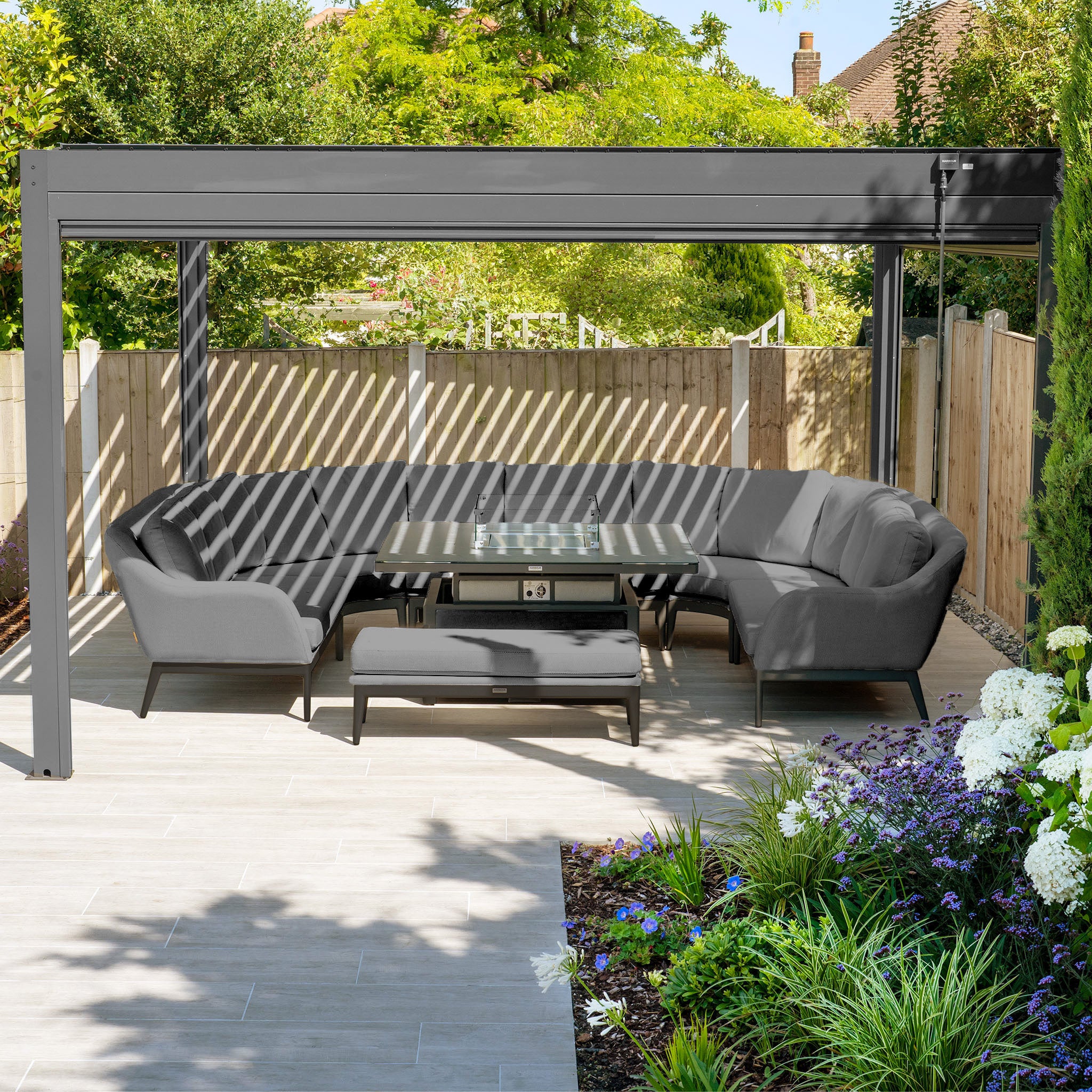 Outdoor patio with a gray Luna U-Shape Outdoor Fabric Corner Dining set, featuring a rising firepit table under a pergola, surrounded by greenery and flowers.