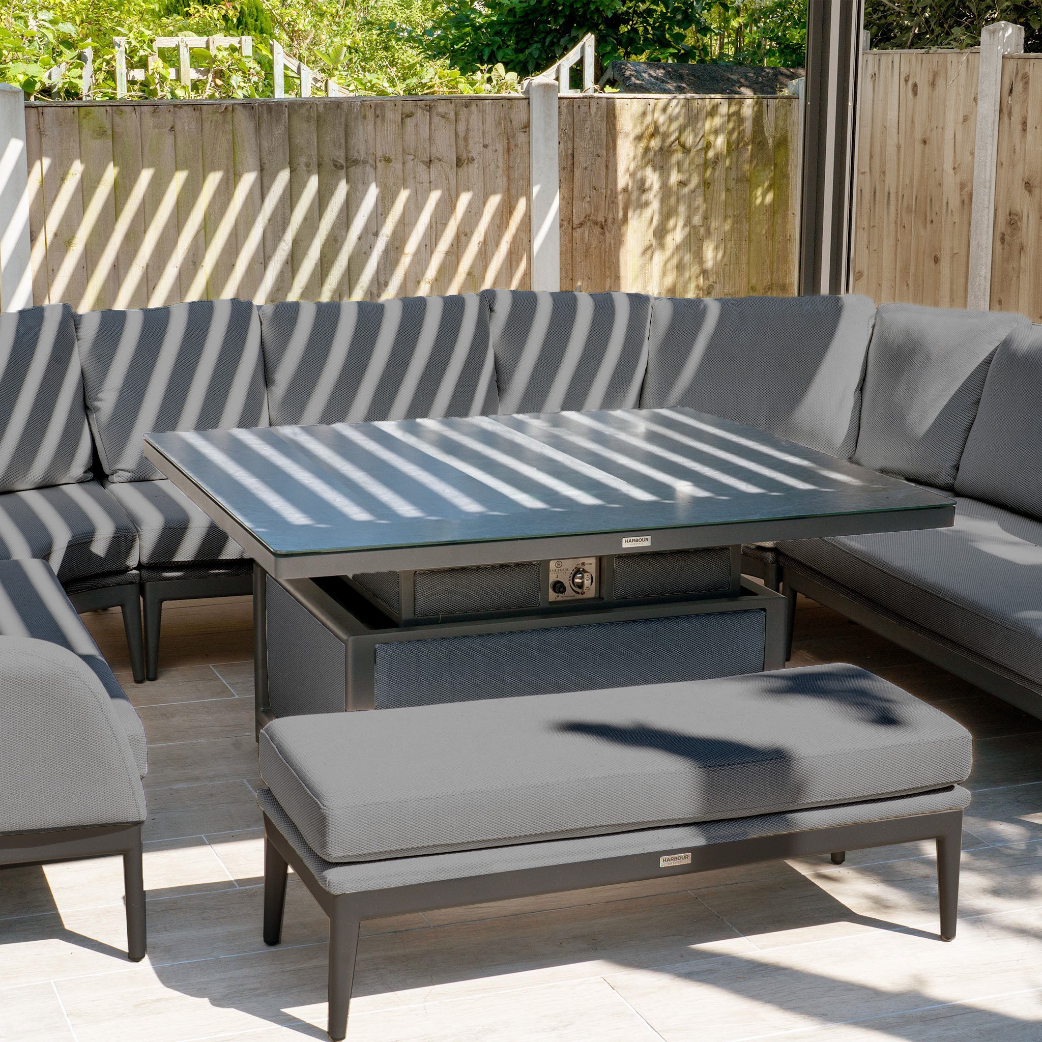 Outdoor seating area with a large square rising table, Luna U-Shape outdoor fabric gray cushioned corner dining set, and a matching cushioned bench.