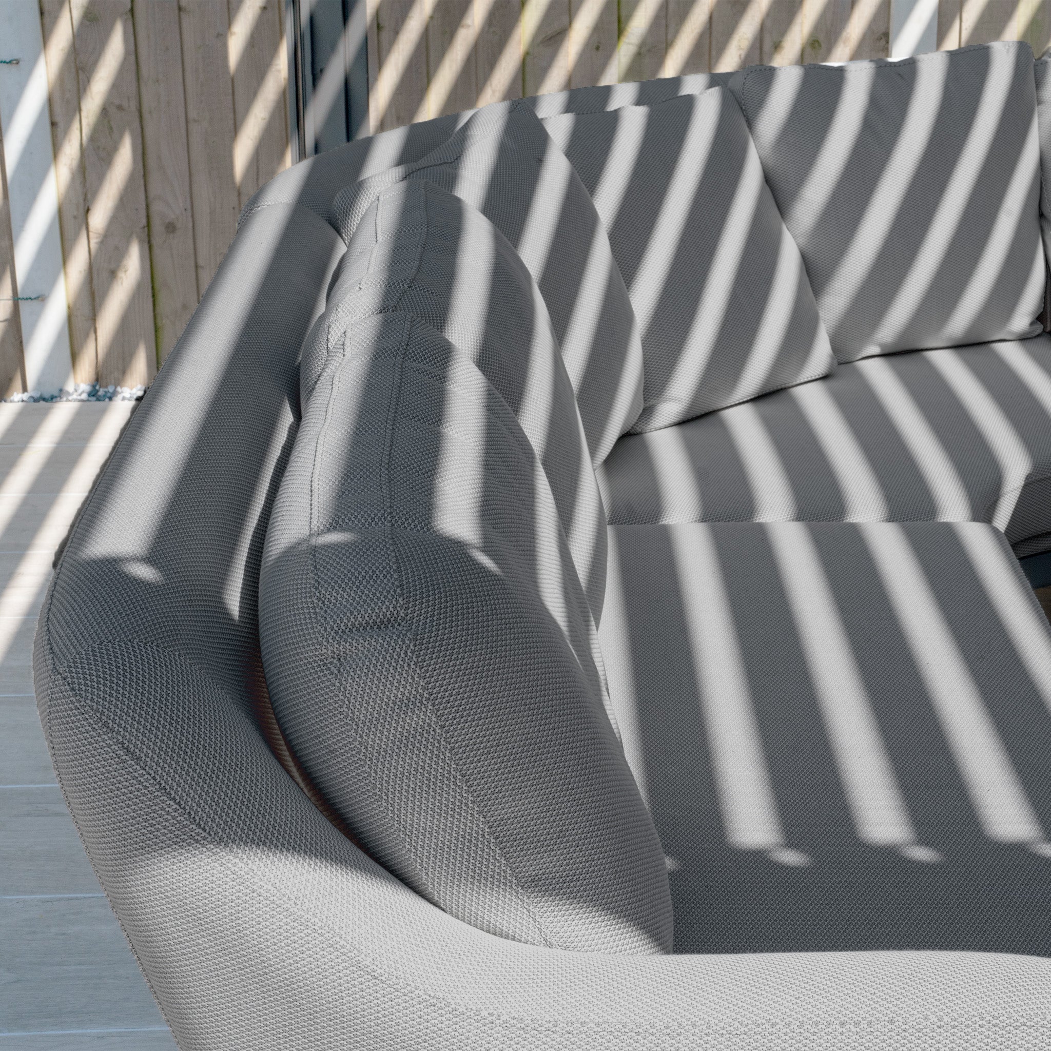 Sunlight filtering through a slatted structure casts striped shadows on the Luna U-Shape Outdoor Fabric Corner Dining set with its grey cushioned curved sofa and Rising Firepit Table.