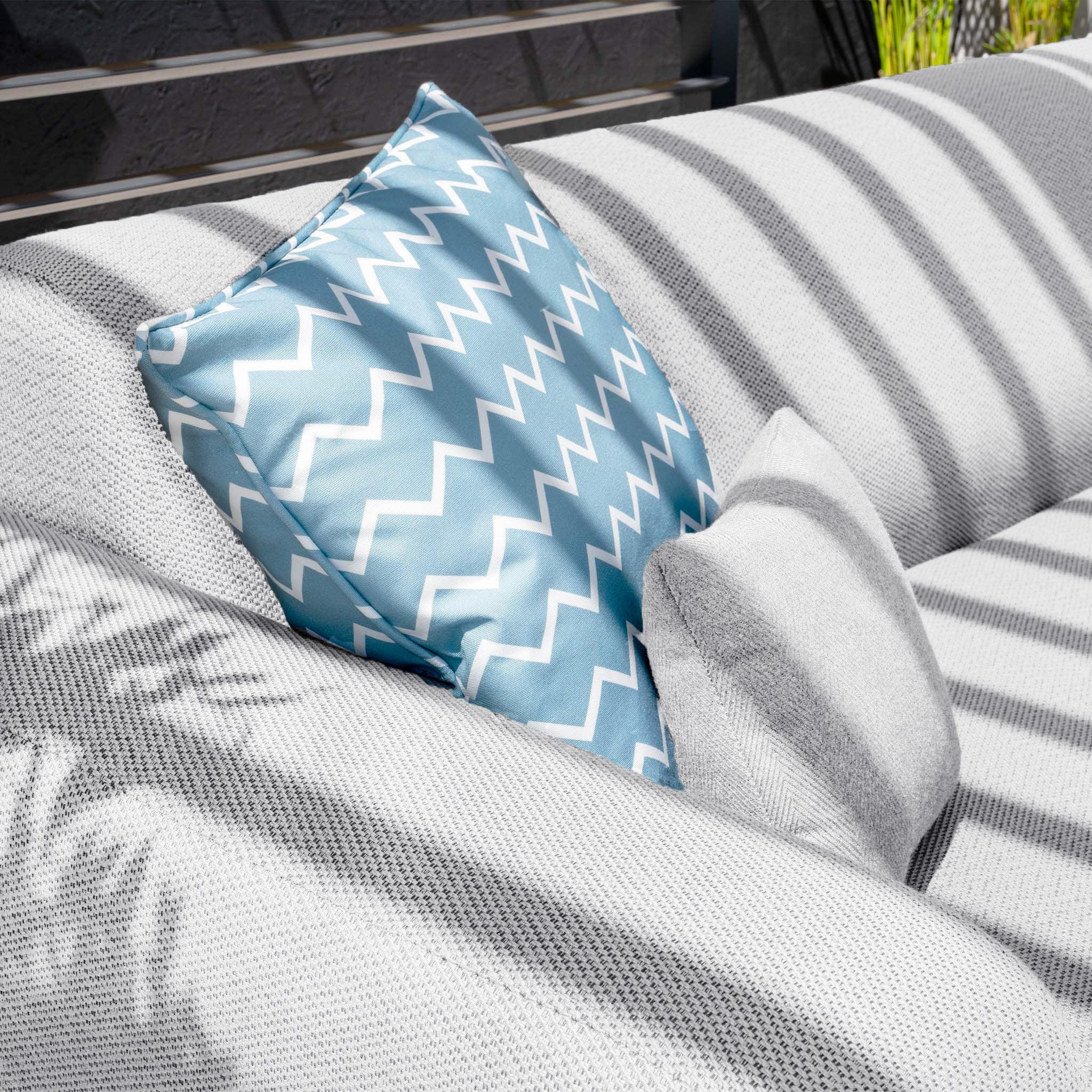 A light blue chevron-patterned pillow and a smaller gray pillow lie on the Luna U-Shape Outdoor Fabric Sofa Set with Firepit Coffee Table in Oyster Grey, basking in the sunlight.