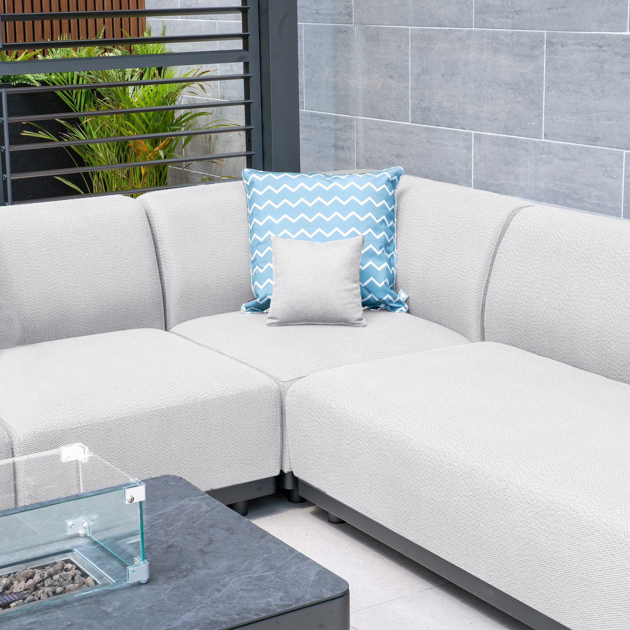 A contemporary outdoor seating arrangement featuring the Luna U-Shape Outdoor Fabric Sofa Set in Oyster Grey, complete with all-weather durability and accompanied by two pillows—one white and one blue—positioned near a glass-top firepit coffee table.