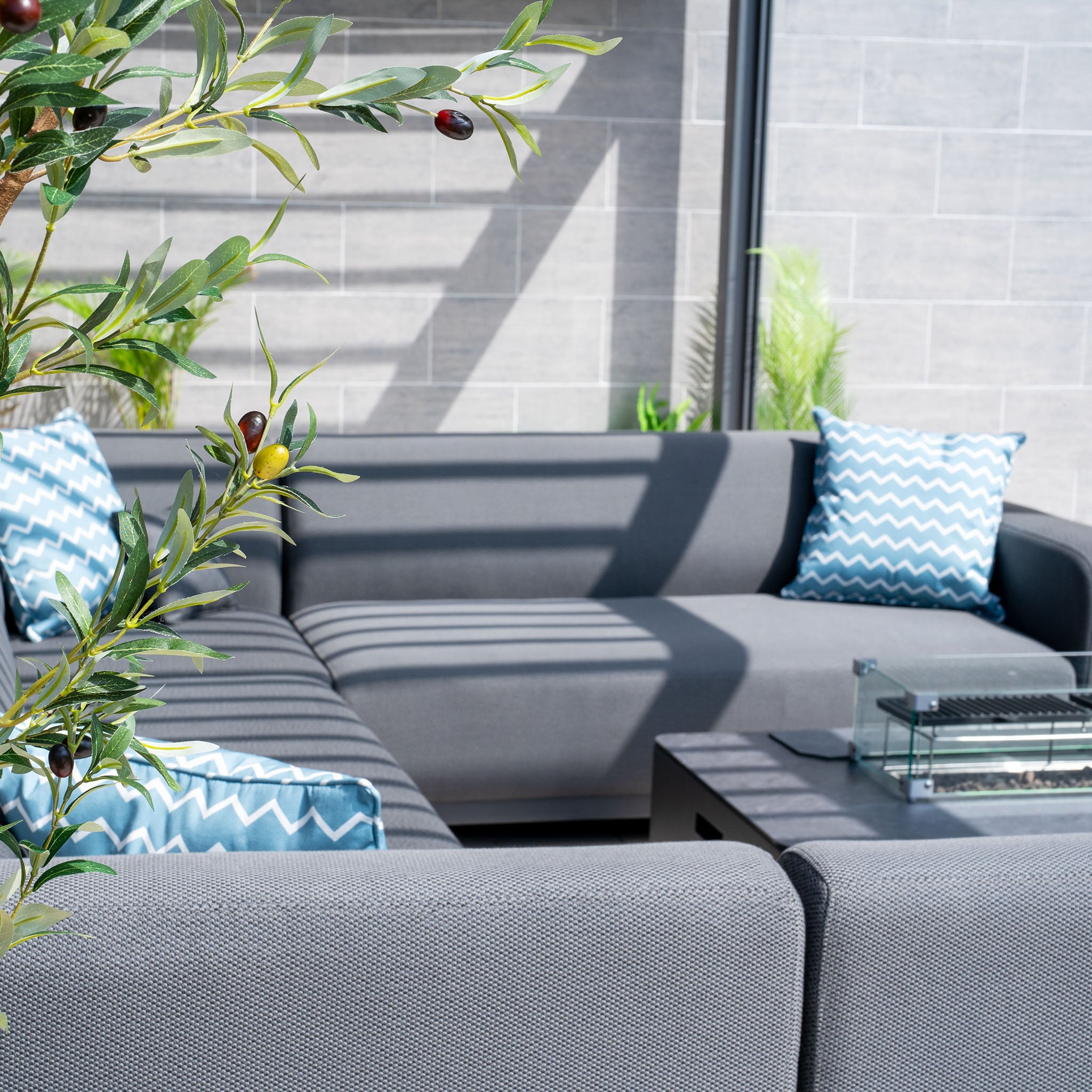 Modern outdoor patio with the Luna U-Shape Outdoor Fabric Sofa Set in gray, adorned with blue patterned pillows and a potted plant in the foreground. A stylish Firepit Coffee Table adds warmth and ambiance to this inviting space.