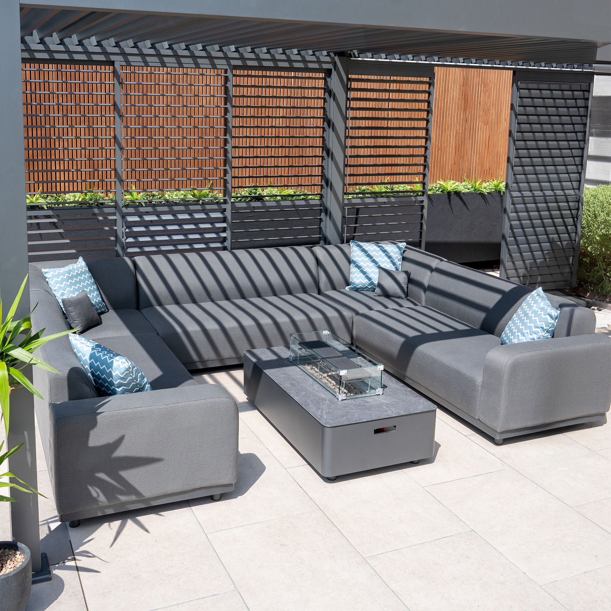 Modern outdoor patio featuring the Luna U-Shape Outdoor Fabric Sofa Set with Firepit Coffee Table in grey, complemented by blue-patterned pillows and a slatted pergola casting shadows.