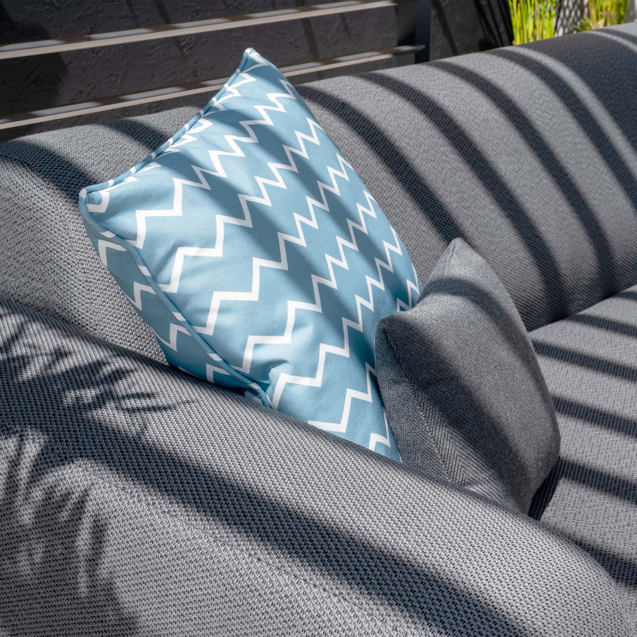 A grey sofa adorned with a teal and white zigzag-patterned cushion alongside a plain grey cushion, all under the dappled light of striped shadows, is perfectly complemented by the nearby Luna U-Shape Outdoor Fabric Sofa Set with Firepit Coffee Table in Grey.