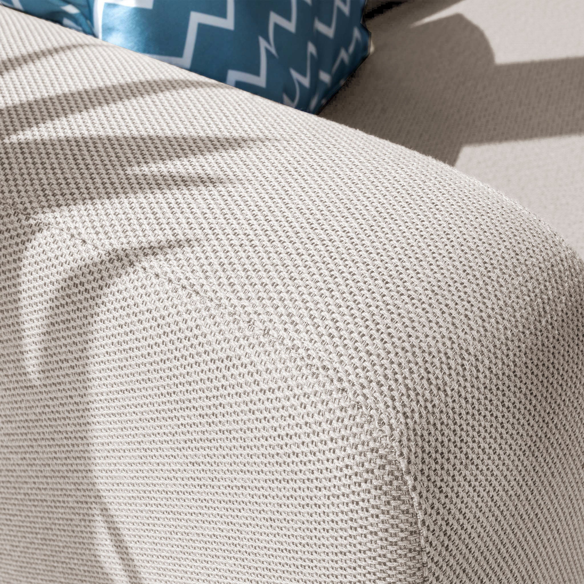 A close-up of the Luna Outdoor Fabric Corner Sofa Set's textured fawn arm, made from all-weather material, with a blue cushion visible in the background.