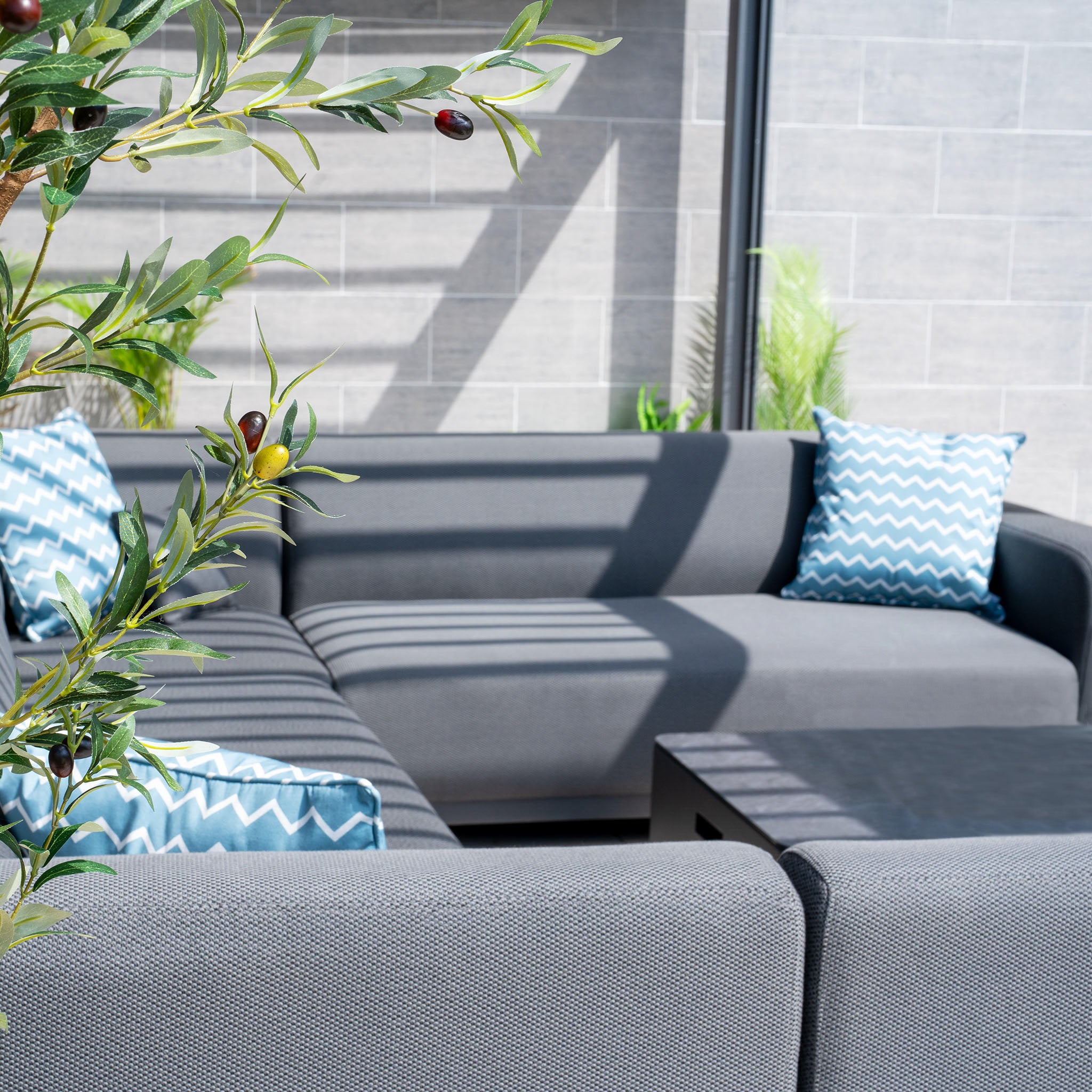 Modern outdoor seating area featuring the Luna U-Shape Outdoor Fabric Sofa Set with Coffee Table in Grey, complemented by blue and white zigzag pillows, and a potted plant.