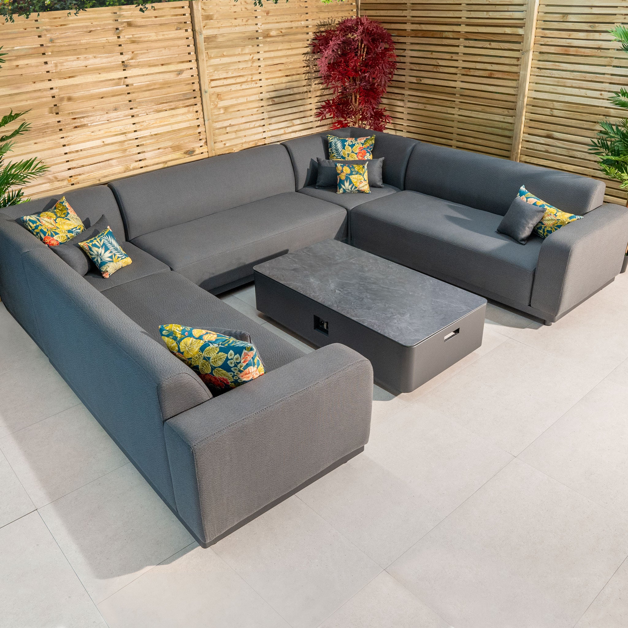 The Luna U-Shape Outdoor Fabric Sofa Set with Coffee Table in Grey features colorful patterned pillows and wooden slat fencing. This maintenance-free sofa set is perfect for any season with its all-weather proof design.