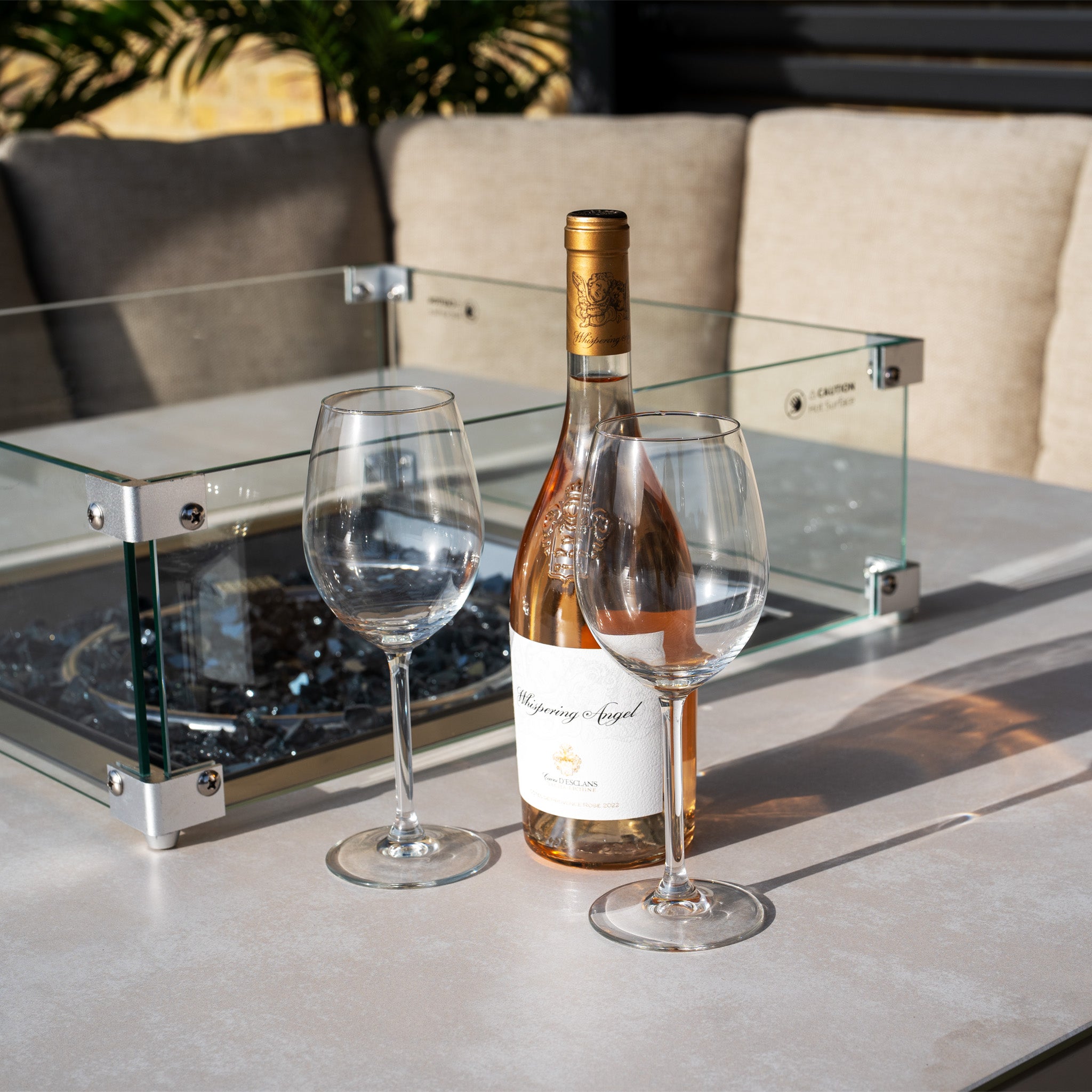 The Luna U-Shape Outdoor Fabric Corner Dining set in Fawn features a rising table topped with a bottle of rosé and two empty glasses, surrounded by cushioned seats. In the backdrop, a cozy fire pit flickers, enhancing the ambiance.