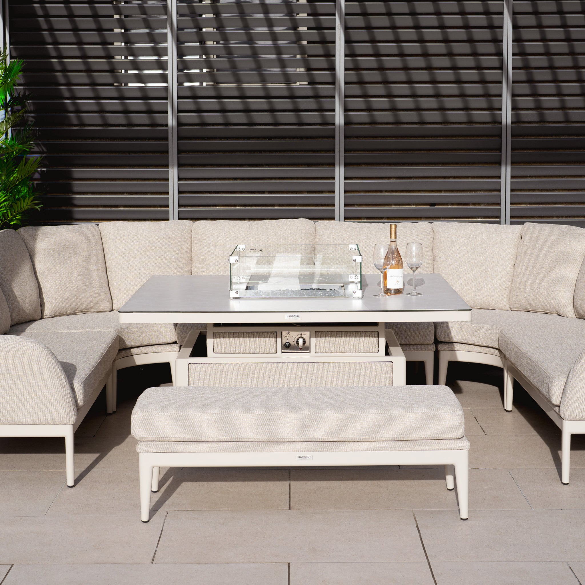 The Luna U-Shape Outdoor Fabric Corner Dining set includes a weather-resistant fawn sofa, a rising table for all occasions, and a cozy fire pit. The wine bottle and glasses on the table add elegance to this modern patio setup.