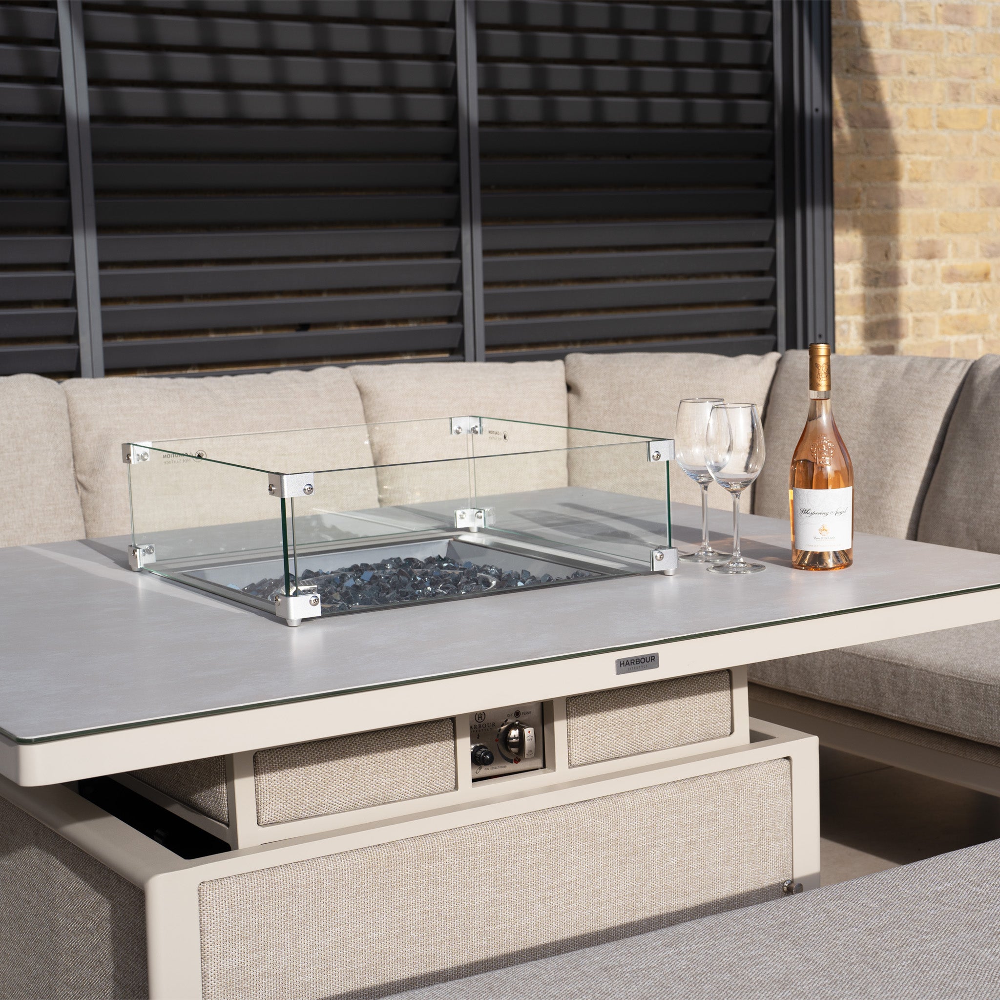 Luna U-Shape Outdoor Fabric Corner Dining with Rising Table in Fawn includes a glass-enclosed fire pit, weather-resistant cushion seating, and a bottle of wine with two glasses.