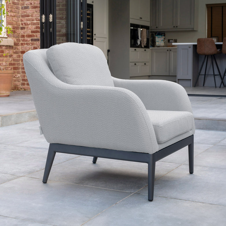 A contemporary Luna Armchair in Oyster Grey from Harbour Lifestyle, featuring sleek metal legs and all-weather outdoor fabric, sits on a tiled patio next to a brick wall and open doors.